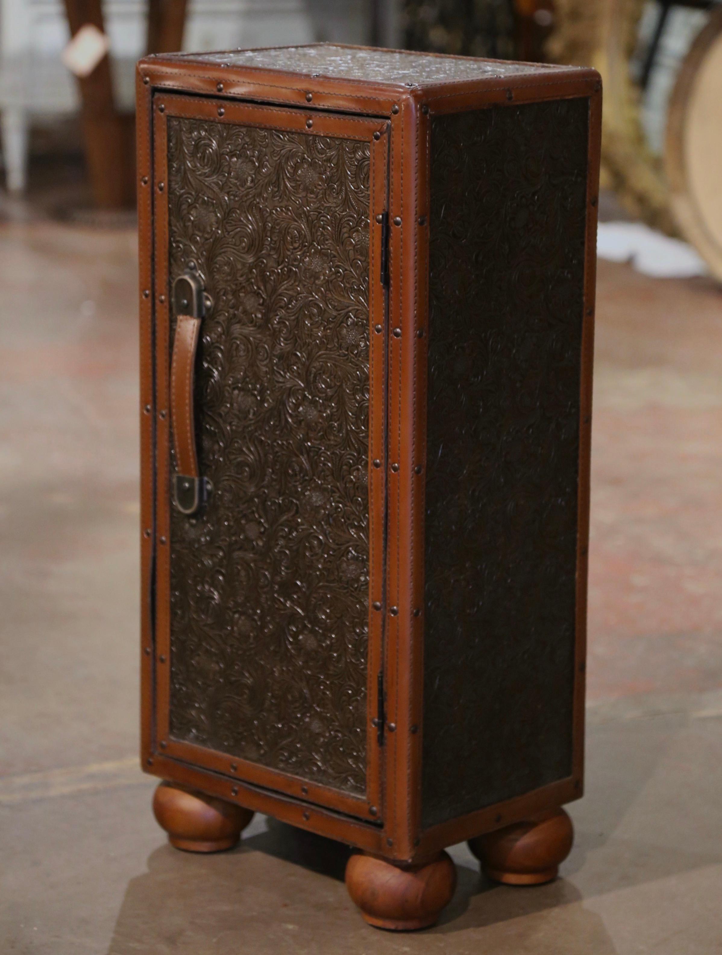 Crafted in Spain circa 1990, the small cabinet stands on bun feet, and is upholstered with a patinated embossed brown leather, embellished with leather straps in each corner. Shaped like a suitcase or trunk, the piece features a center door dressed