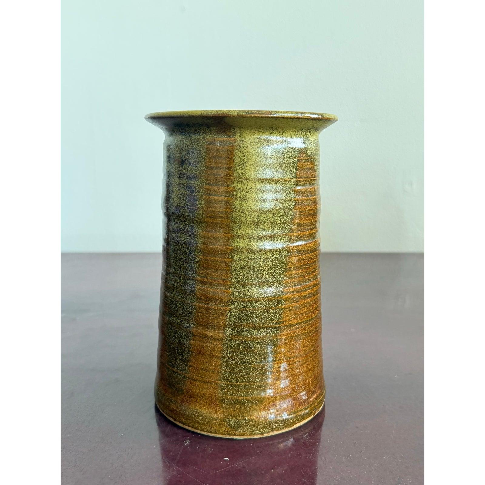 Esta jarra de cerámica labrada a mano presenta un rico vidriado que varía entre verdes terrosos y marrones óxido intenso, con sutiles abigarramientos y crestas de textura natural procedentes del torno. El labio acampanado le da una forma clásica,