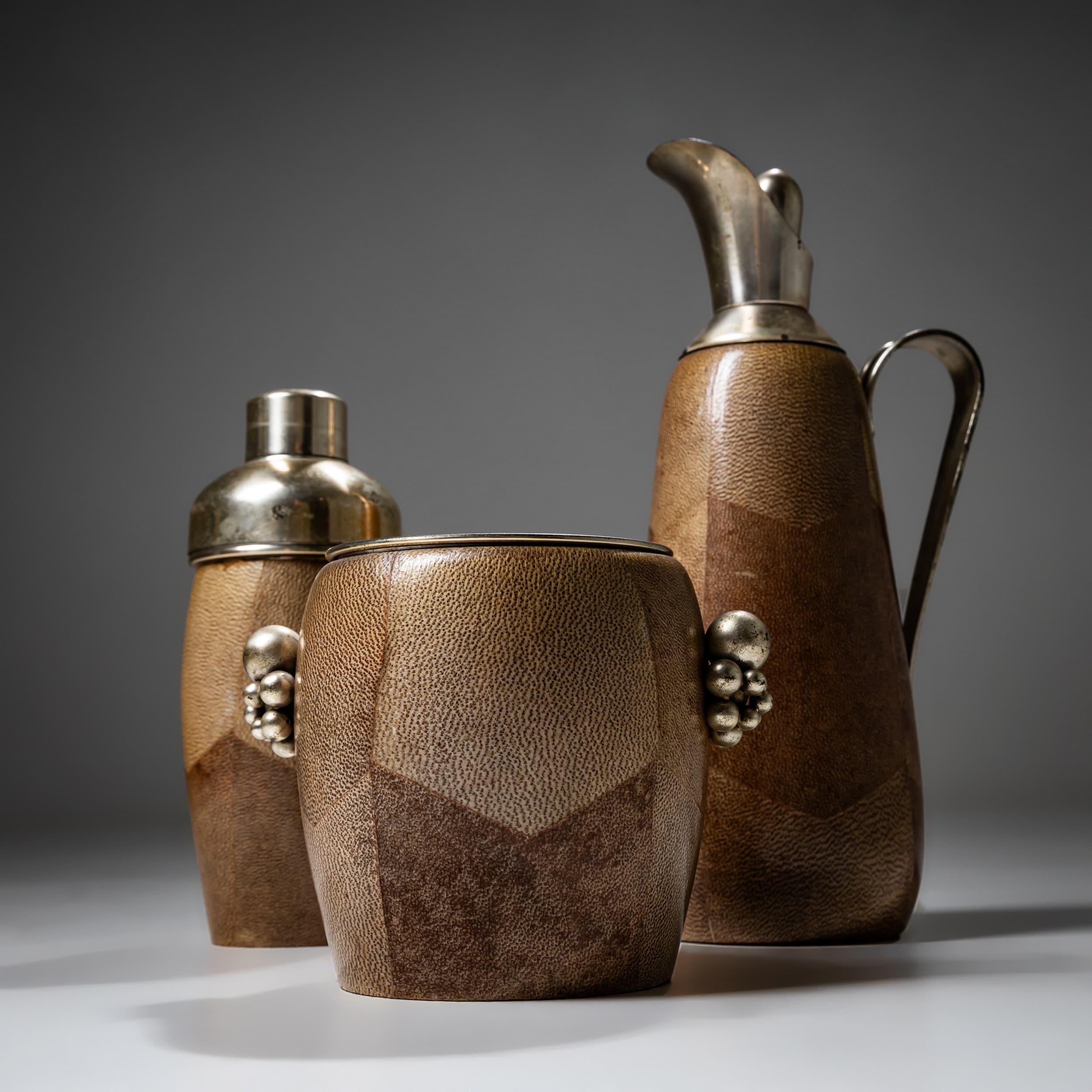 Elegant three-piece bar set by Aldo Tura, comprising a tall carafe (12.2 x 5.9 x 4.7 in), a cocktail shaker (8.7 x 3.5 in), and an ice bucket (5.3 x 6.7 x 5.1 in). Each piece is covered in tobacco-colored goatskin and finished with clear lacquer.
