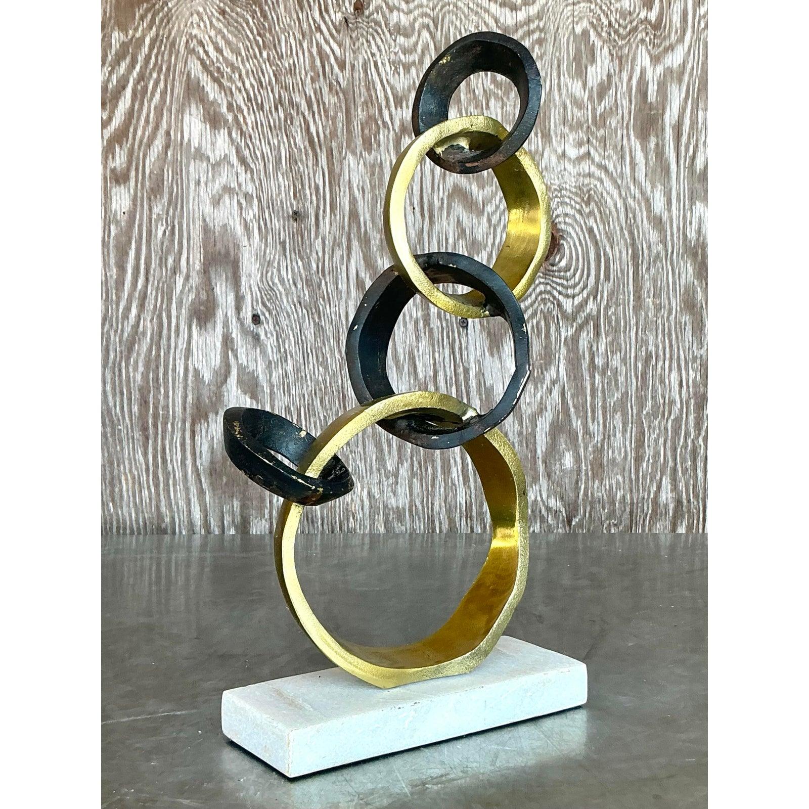 Eleva i tuoi interni con questa sorprendente scultura astratta vintage caratterizzata da una catena ritmica di anelli intrecciati in metallo annerito e dorato. Appoggiato ad arte su una base di marmo bianco, questo pezzo bilancia il fascino
