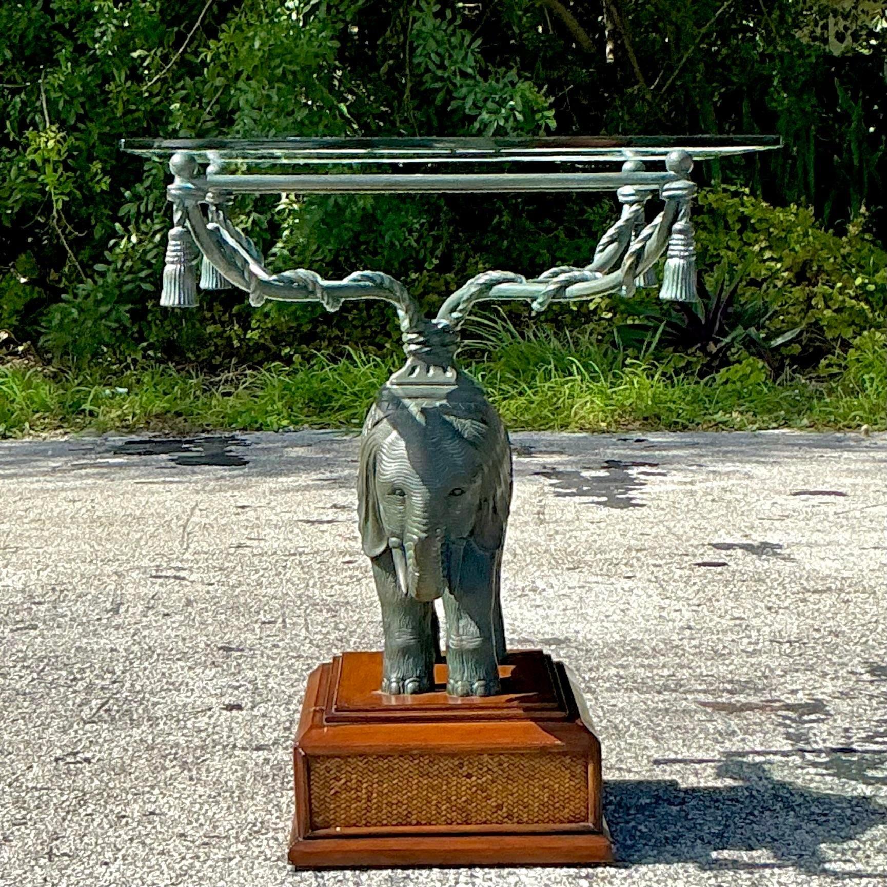 This unique vintage side table features a beautifully patinated elephant base, lending an exotic and bohemian touch to any room. The elephant sculpture stands proudly on a square wooden base with a woven rattan accent, supporting a clear glass top