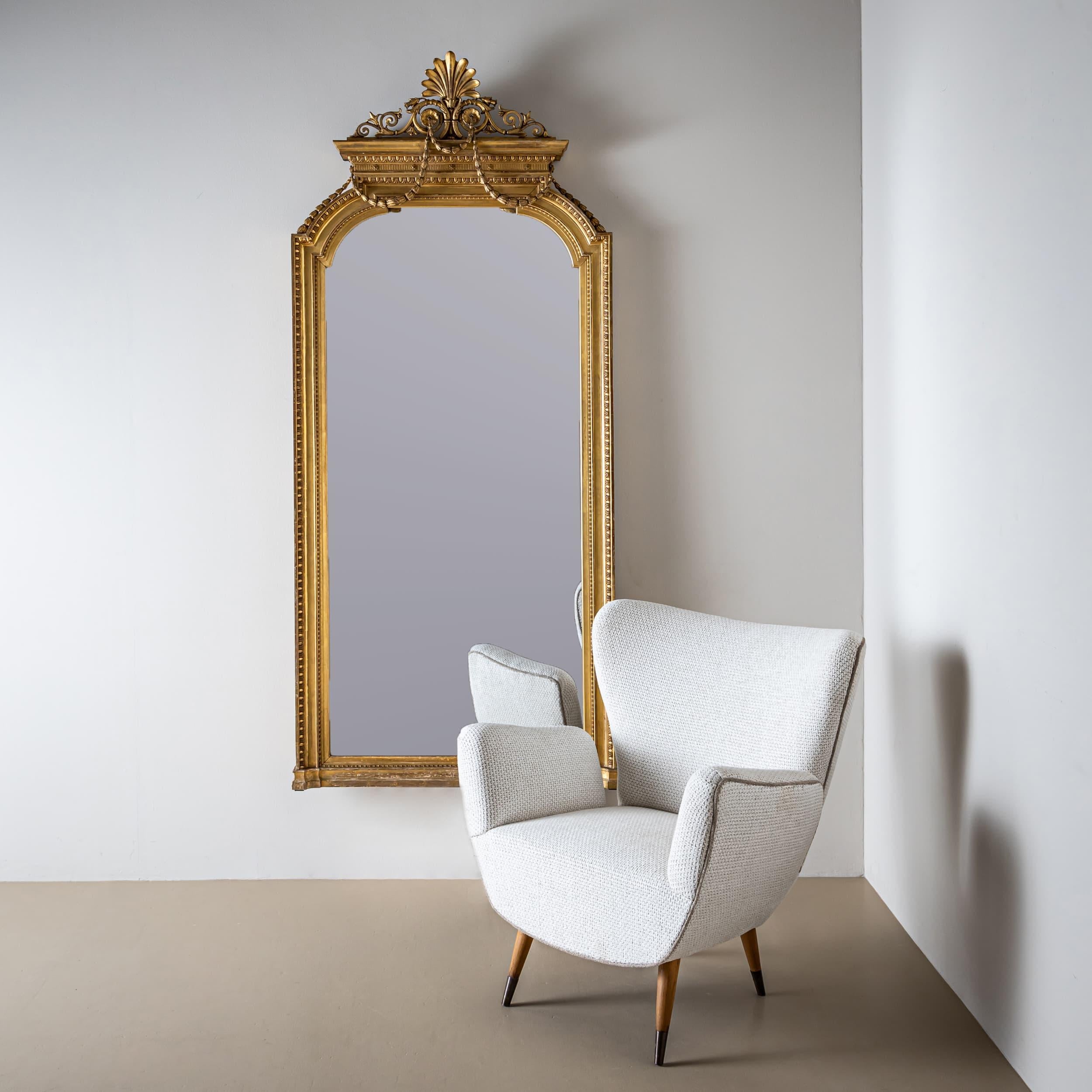 Large and ornate wall mirror from the second half of the 19th century, crafted in the Louis XVI style. The gilded frame features refined beading and bull’s-eye motifs, lending the piece a timeless elegance. The rounded pediment is crowned with an