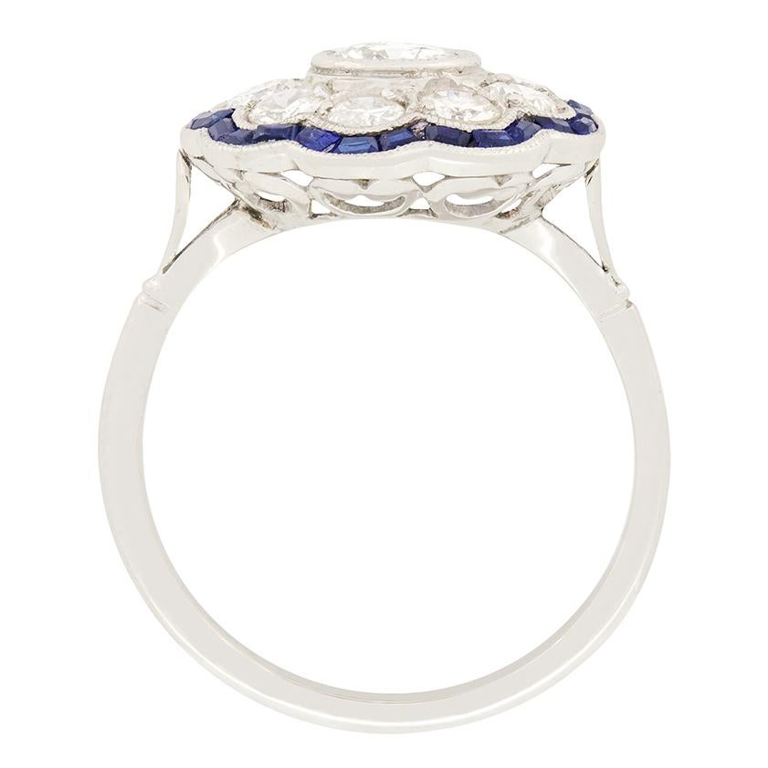 This stunning late deco ring features a total of ten transitional cut diamonds along encircled by a ring of sapphires. The central diamond is 0.65 carat with the surrounding nine weighing a further 0.15 carat each. All the stones are graded G to H
