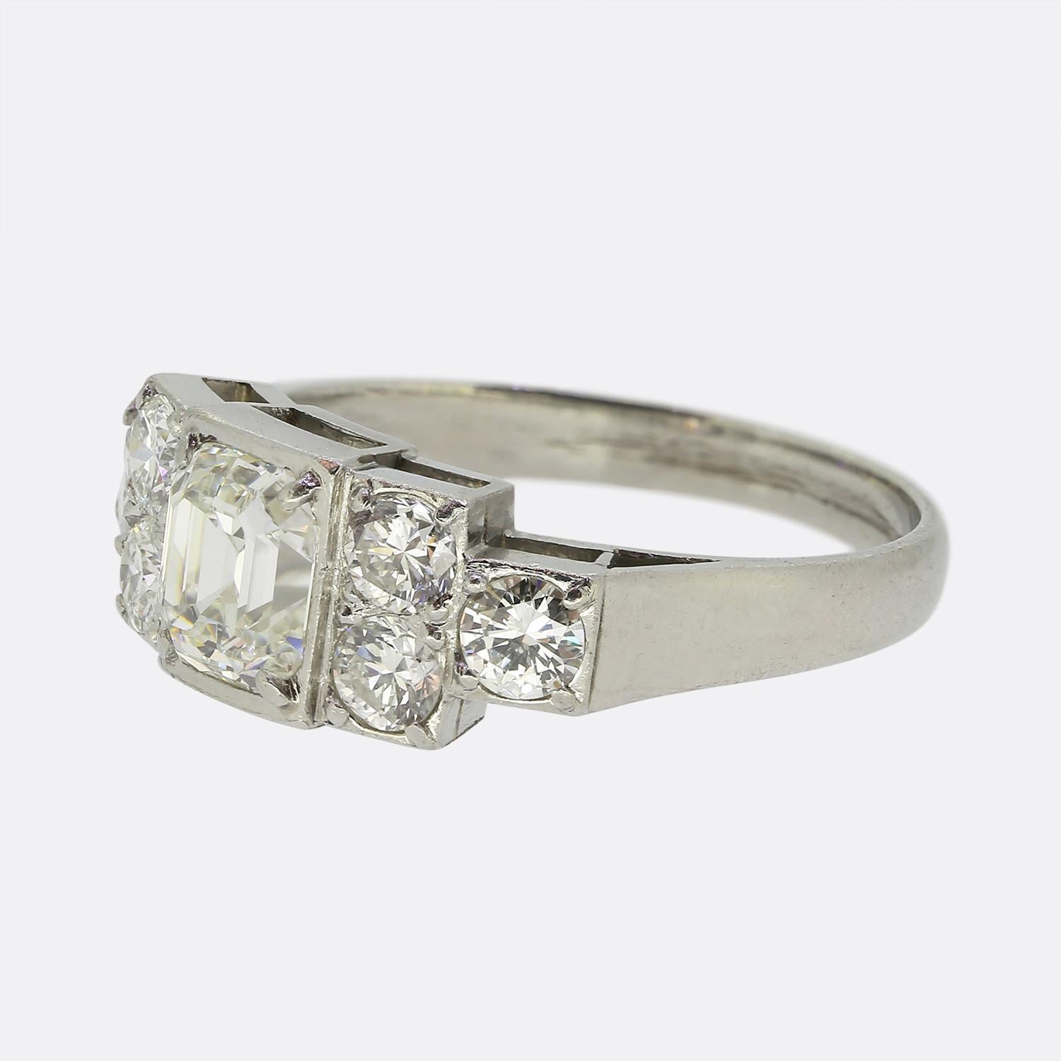 Here we have a striking diamond ring crafted during the 1940s. This platinum piece features a centralised asscher-cut diamond at the centre of the face which sits slightly risen in four-clawed setting. Flanking this principal stone on either side is