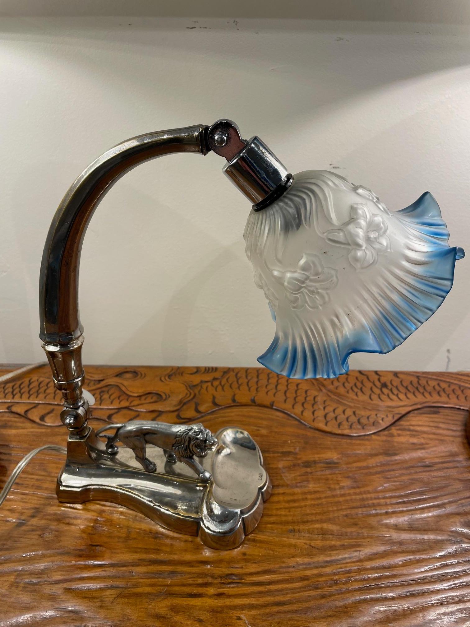 From the late Art Deco period, a table lamp with a silver-plated base depicting a stalking lion.  The shade is a blue and frosted art glass and is adjustable at the arm just above the shade, and also adjusts at the bottom portion of the arm. 