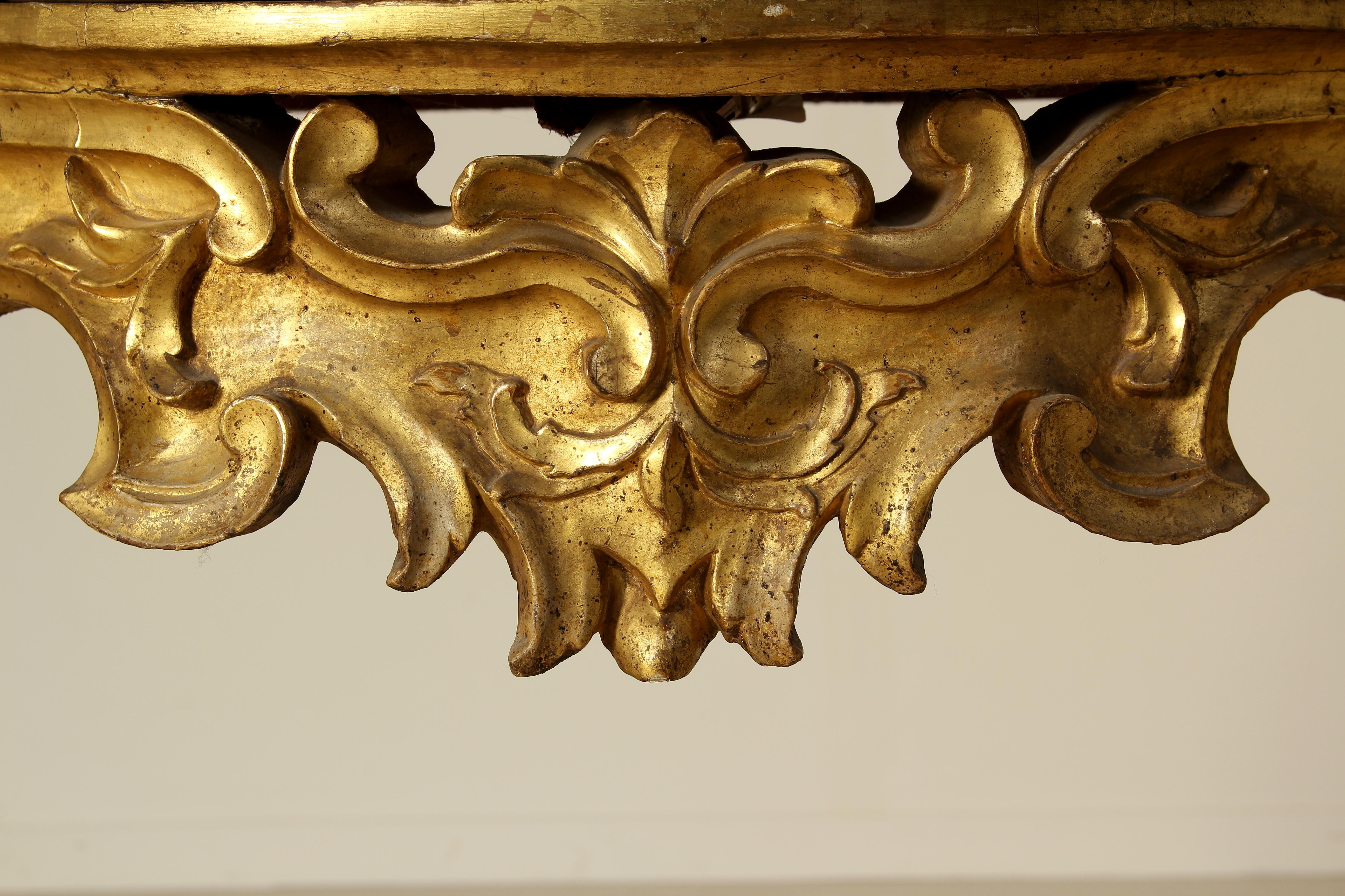 A late Baroque wall table with four carved uprights shaped like goat legs linked with a carved cross centered with a shouting mask under the top. Carved volutes on the front and the sides. The mixtilinear top broadens toward the bottom where the