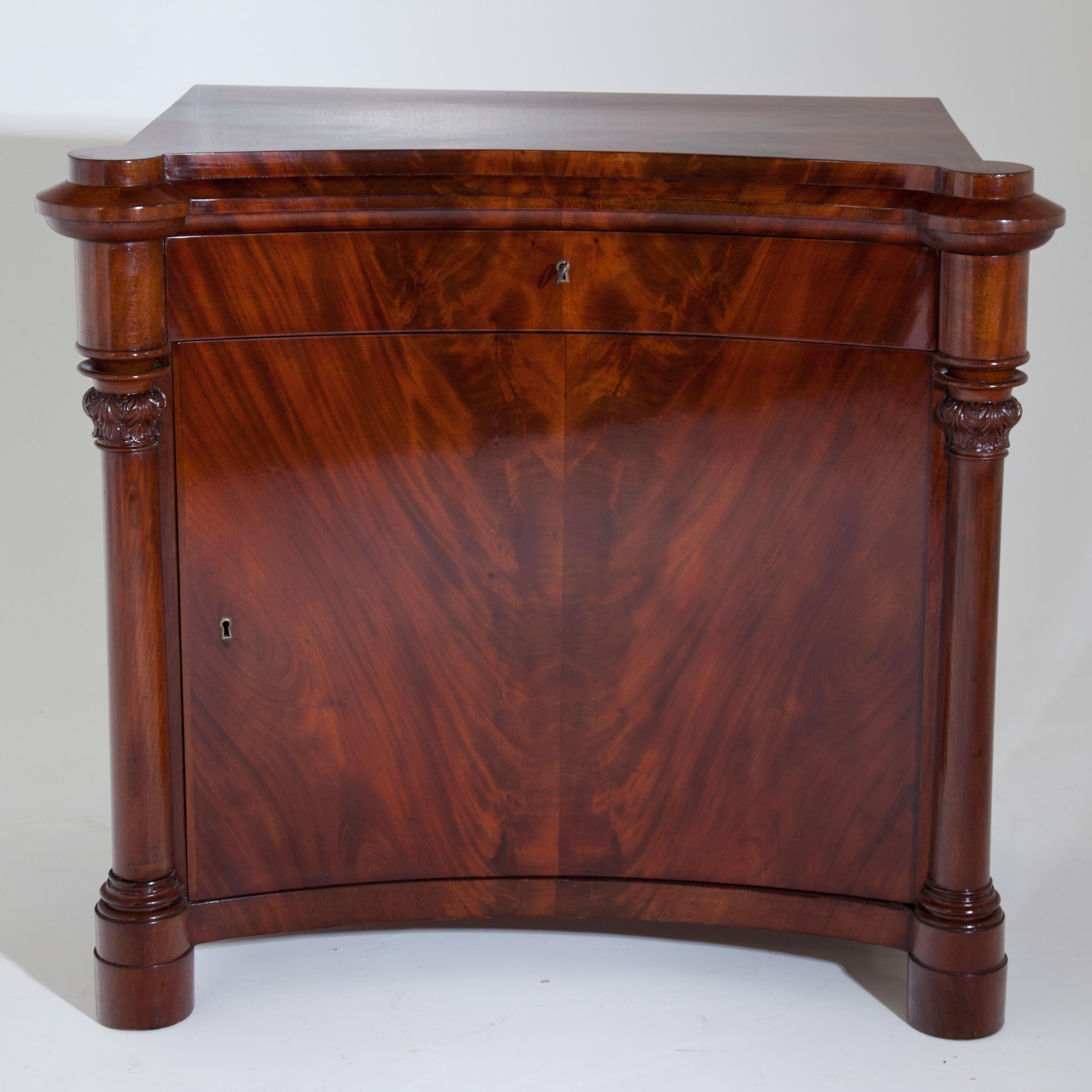 Cabinet with concave curved front and lateral three-quarter-columns with leaf capitals and profiled bases. One-drawer and one-door under the profiled top panel. Mahogany, solid and veneered.
        