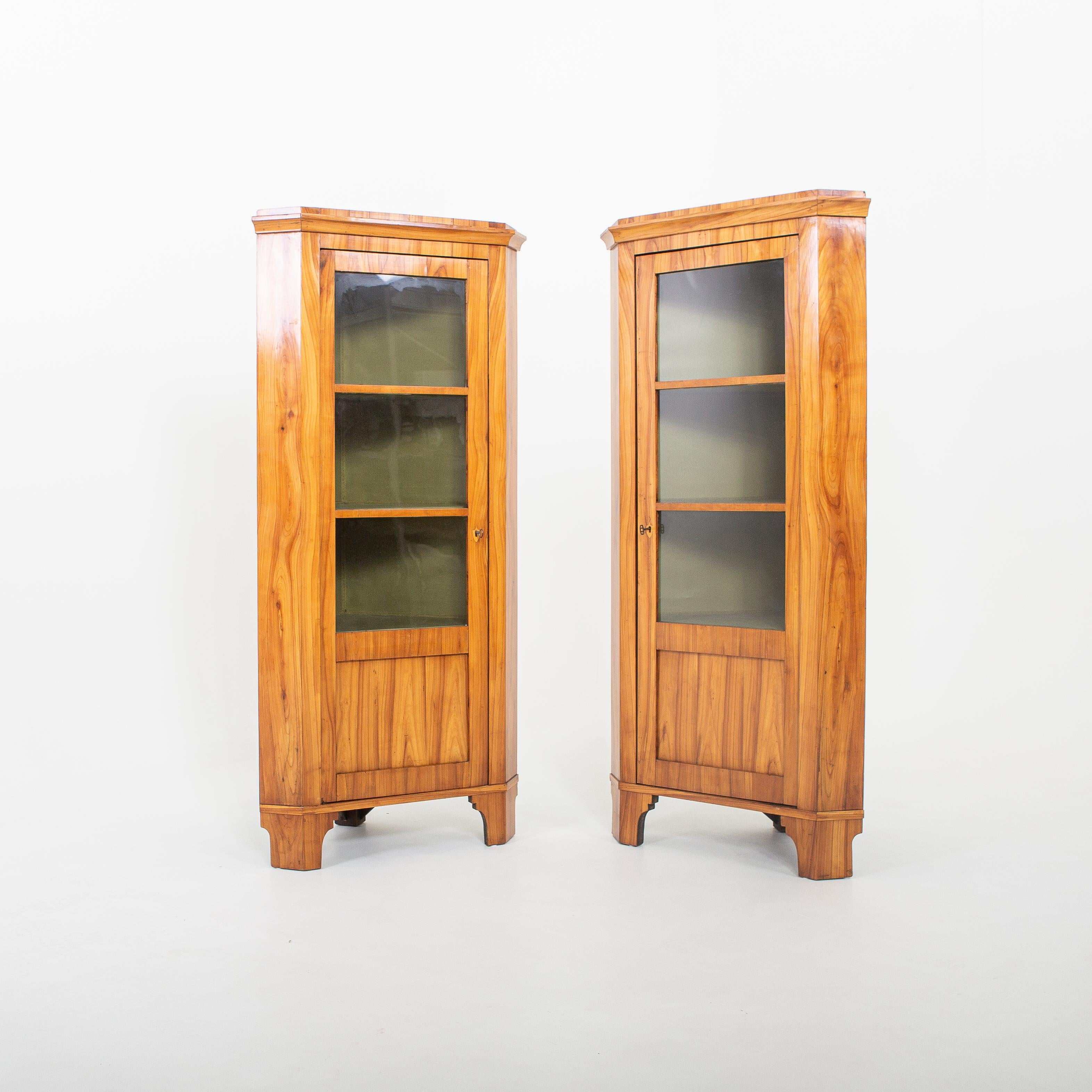 Pair of Biedermeier corner cupboards on bracket feet and a door with three-quarter glazing. Cherry veneered, inside lined with green fabric. Restored condition.