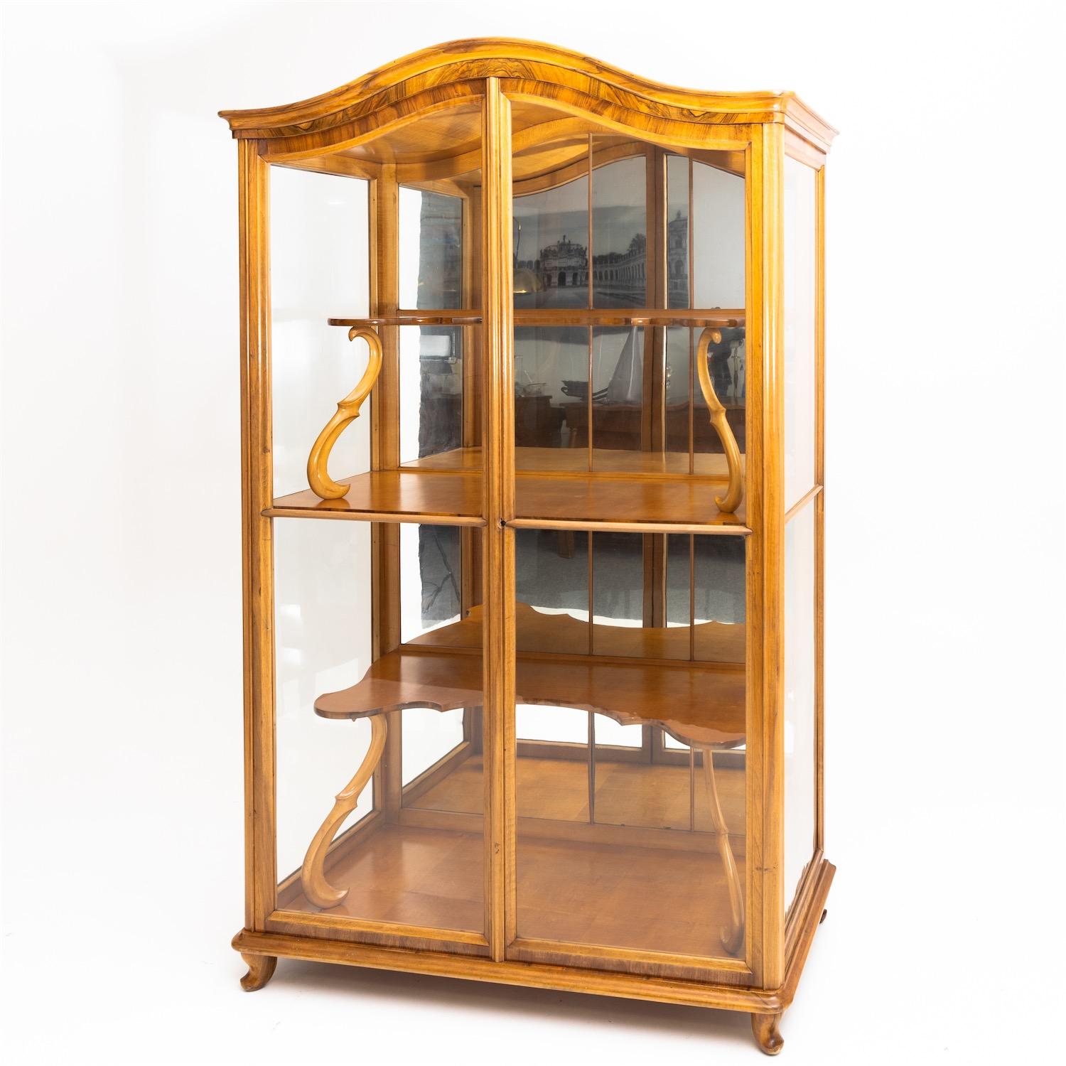 Late Biedermeier display case glazed on three sides with a curved pediment standing on low curved feet. The interior division consists of a full-length shelf and f-shaped supports with curved cut-out recessed shelves. The back is mirrored. Walnut,