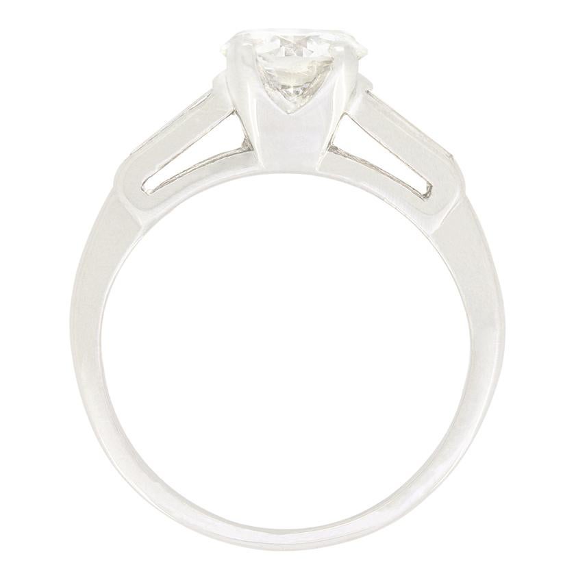 Taking centre stage in this beautiful Late Deco solitaire ring is a 0.85 carat, transitional cut diamond. The diamond has been estimated as I in colour and SI2 in clarity. Flanking the show piece stone, a pair of tapered baguette cut diamonds
