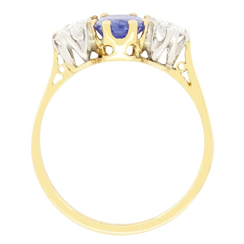 This elegant  1940s three stone ring features a captivating Ceylon sapphire at its centre. The cornflower blue sapphire is an unheated natural stone weighing 1.25 carats and claw set within an 18 carat yellow gold collet. Flanking the sapphire are