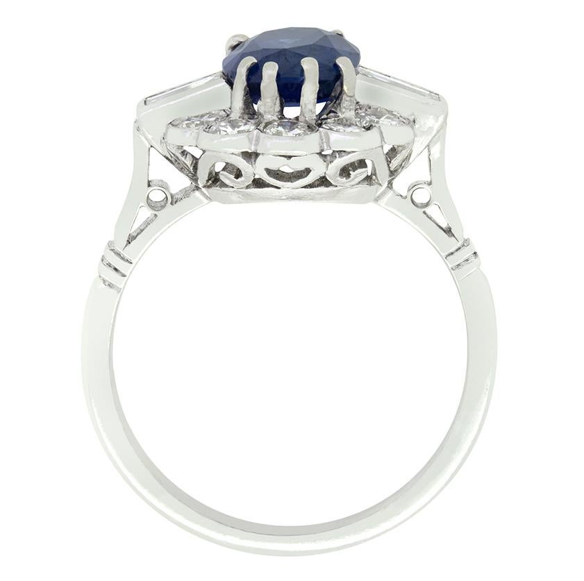 An impressive 1.90 carat sapphire sits enveloped in a cluster of round brilliant diamonds in this Late Deco cluster ring. The oval cut sapphire has a beautiful deep blue tone which, set amongst the whiteness of the diamond contrasts beautifully.