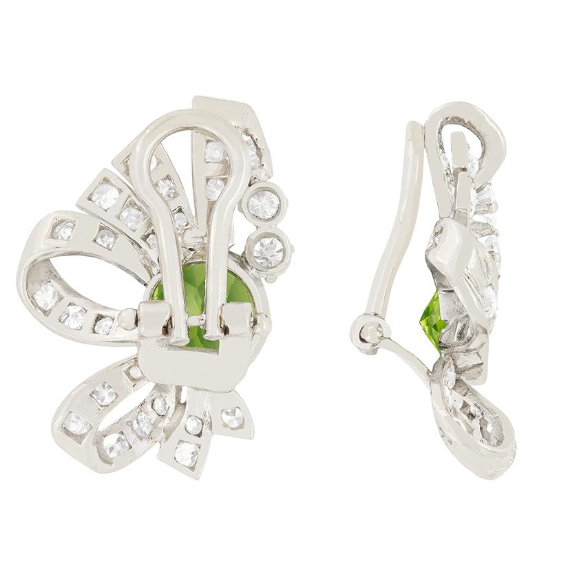 These captivating earrings date back to the brilliant Late Deco era and feature two gorgeous peridot gemstones. The two stones weigh 1.50 carat each and have been rub over set into platinum settings. Surrounding the peridots are a cluster of
