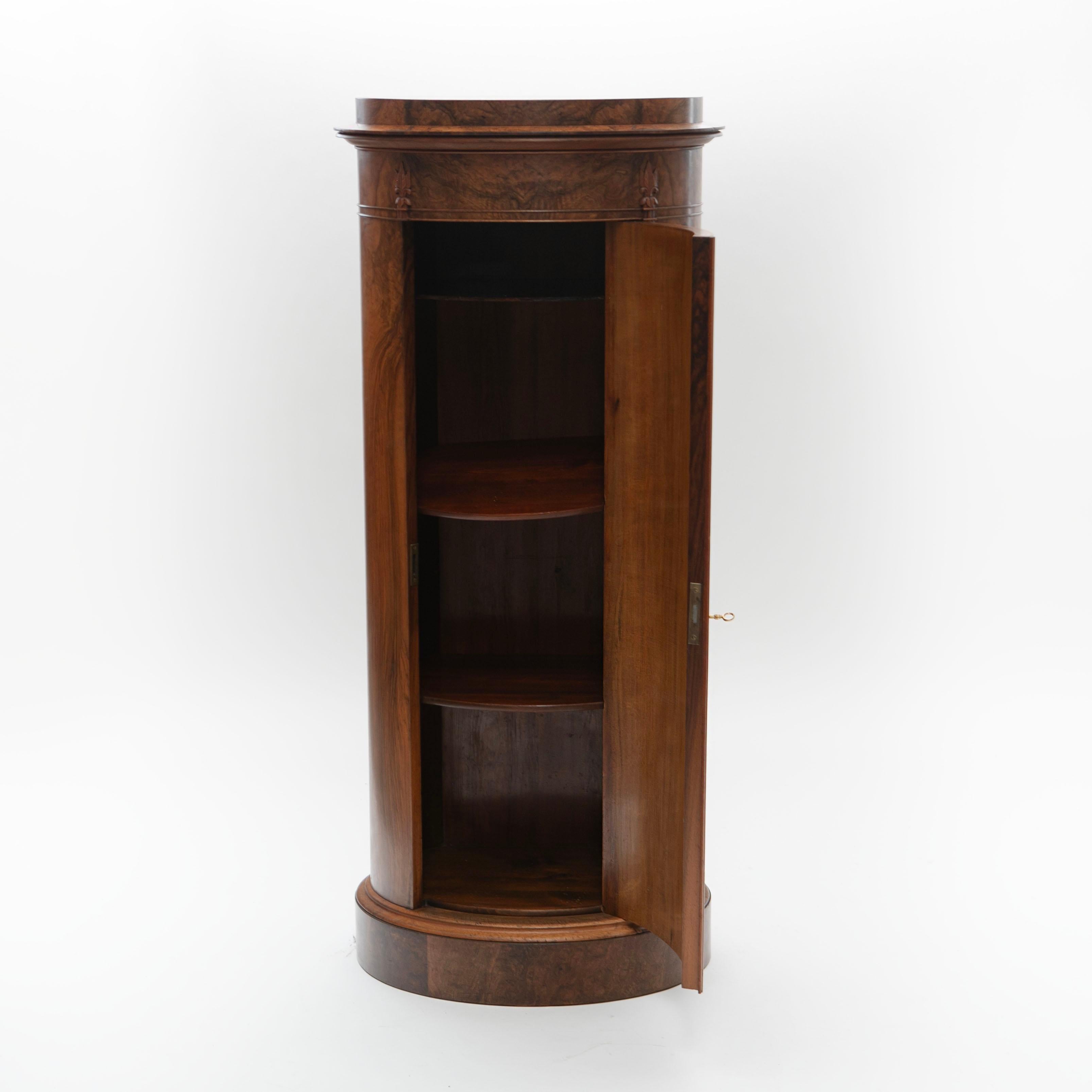 An oval pedestal cabinet veneered in striking walnut root. The front features a single door, revealing shelves inside, while fluted profiled moldings adorn the sides.
The top is crowned with hand-carved acanthus leaves and foliate details.
This
