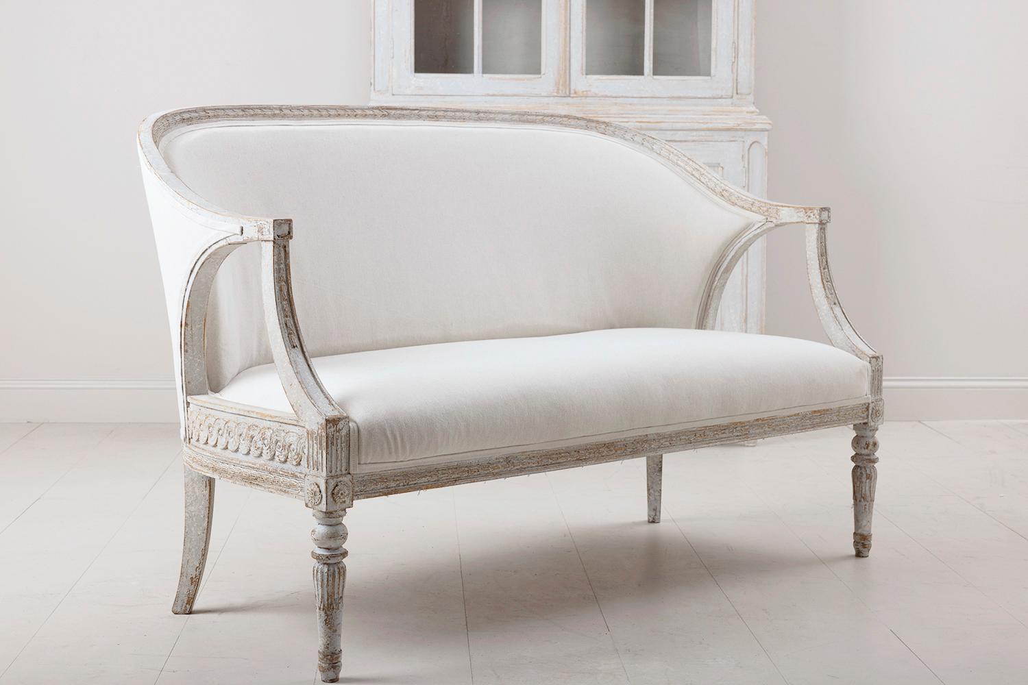 A stunning Swedish sofa in the Gustavian style in original paint with a comfortable barrel-style back, newly upholstered in linen, circa 1900. The frame features carved bell flowers along the front and a carved guilloche pattern on the sides while