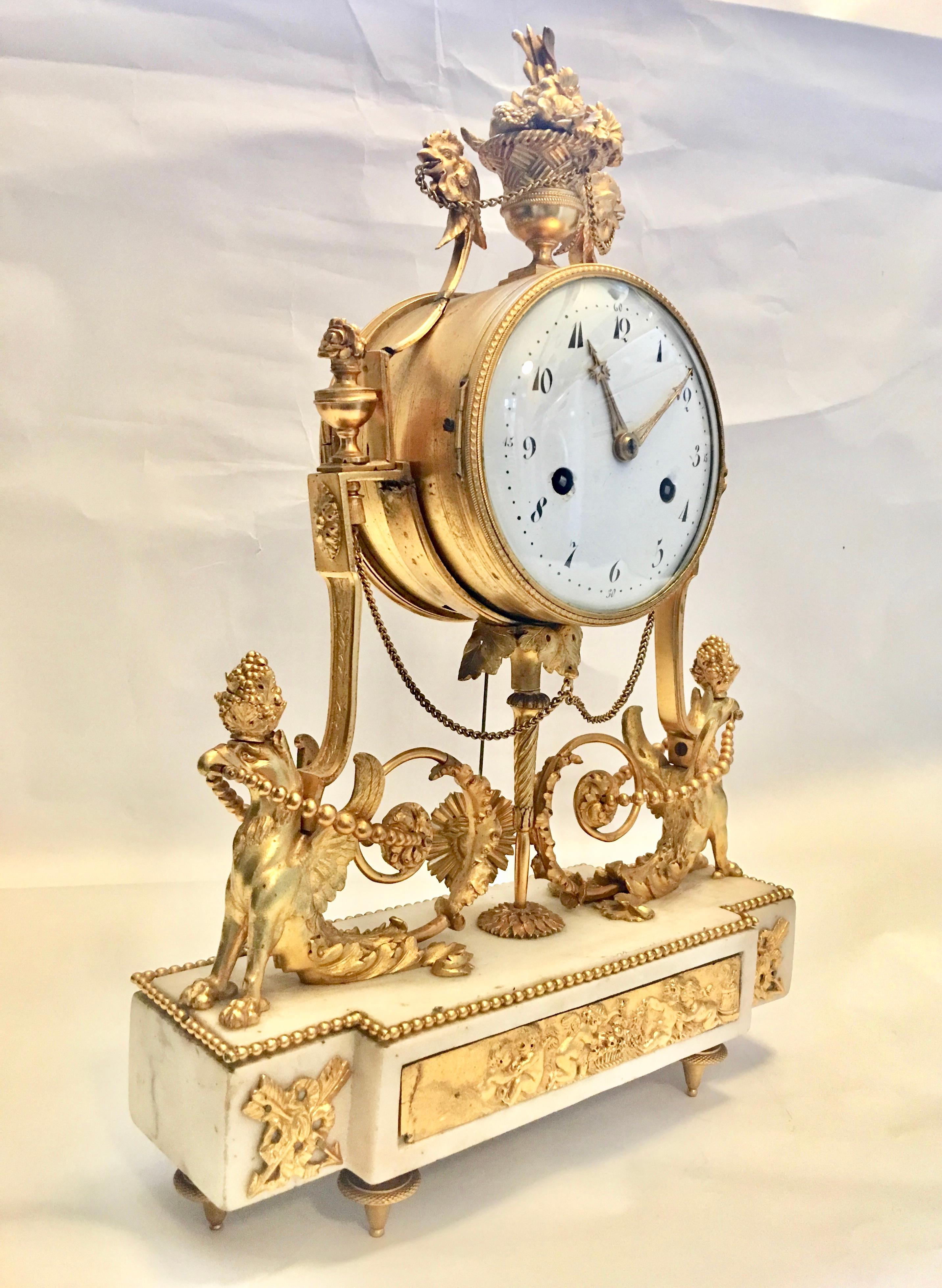 A very fine Piece of Late Louis XVI ormolu and white marble mantel clock,
circa 1800.

The drum-shaped case surmounted by a flower-filled basket flanked to each side by a cockerel head, on strapwork frame with a pair of seated chimera supports