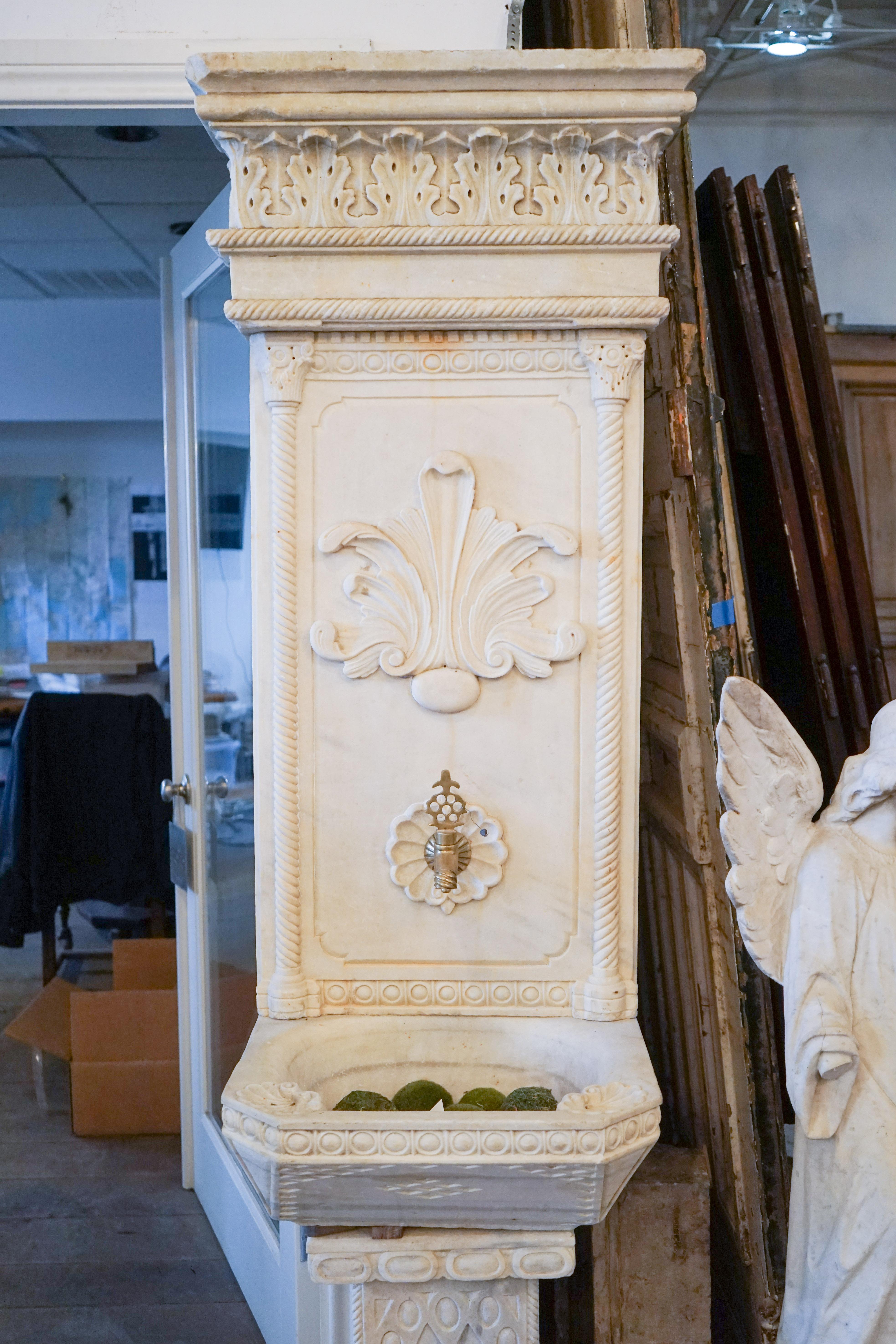 Seated on a carved square pediment, this creamy ivory marble wall fountain features spiral inset columns and molding. A large classical anthemion motif dominates the back splash and repeats atop the cornice. The basin features finely carved