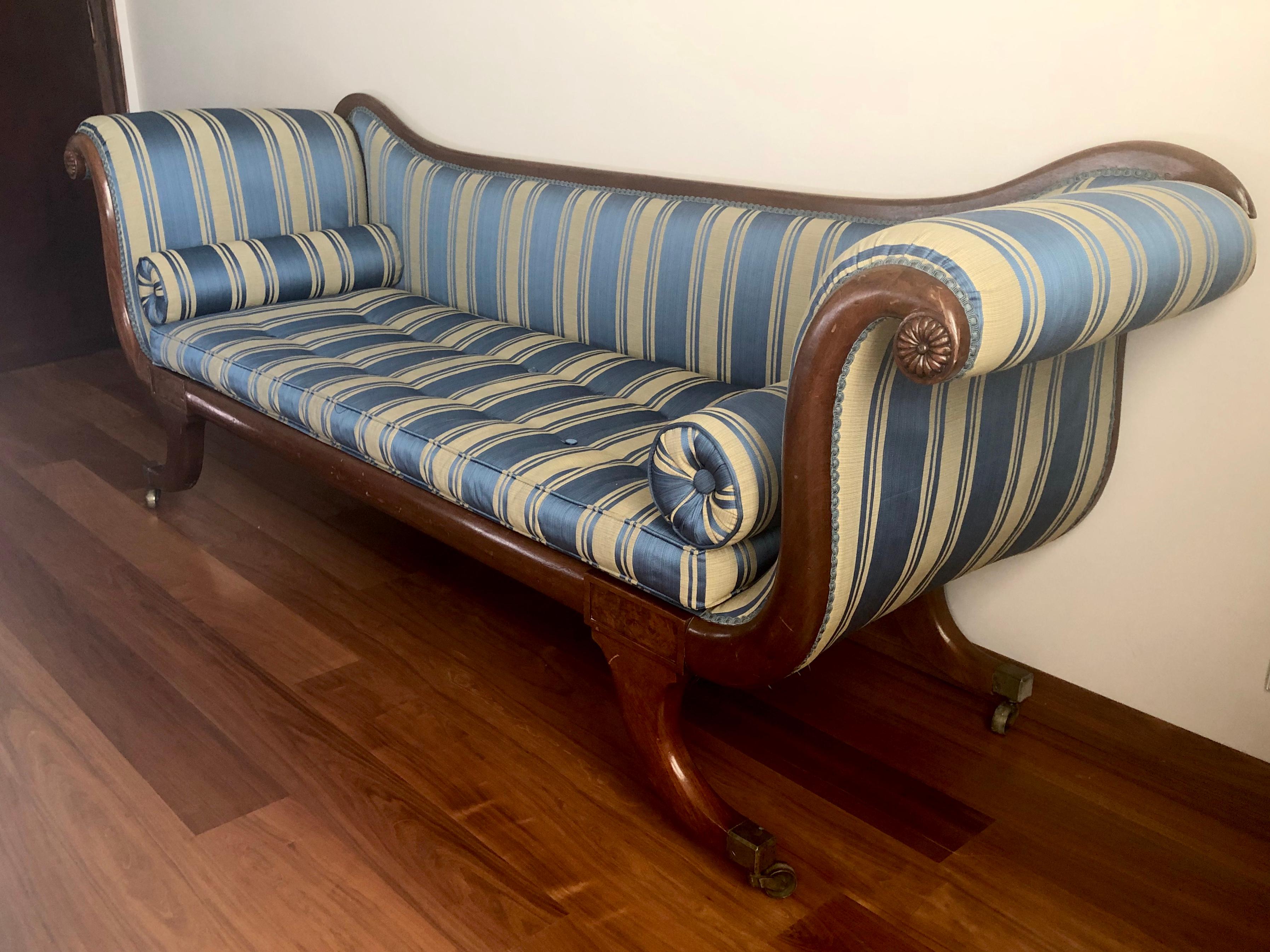 A late Regency mahogany scroll end sofa, with a detachable back, with blue striped fabric, squab cushion and pair of bolsters, the frame applied with lobed rondels and flame veneered tablets, on brass castors.
England, circa 1820.

 