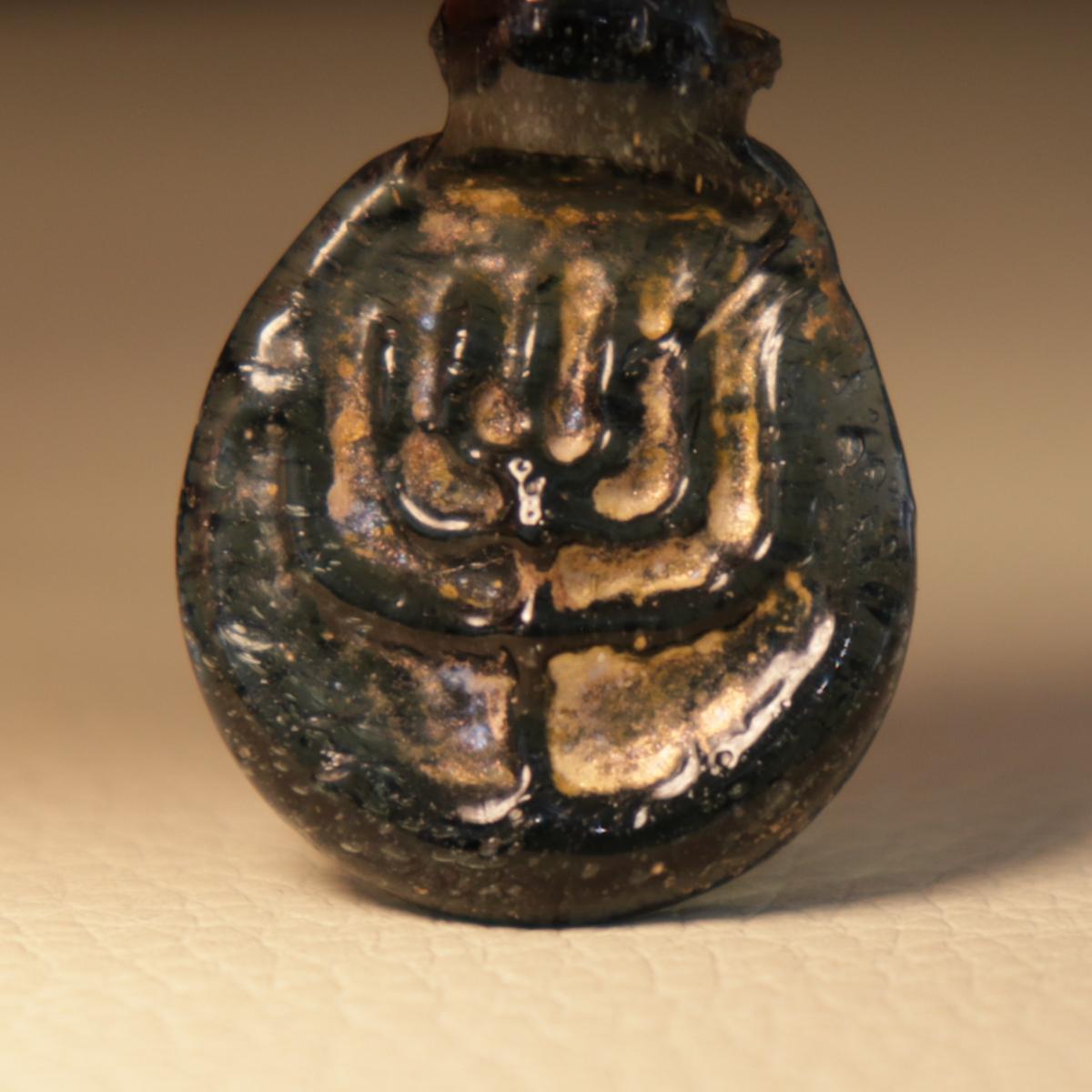 Late Roman to Early Byzantine, ca. 300 to 500 CE. A rare and beautiful amulet of a generally circular form with a flat bottom, a raised periphery, and an integral suspension loop, mold-pressed from attractive cobalt blue glass to depict a seven-lamp