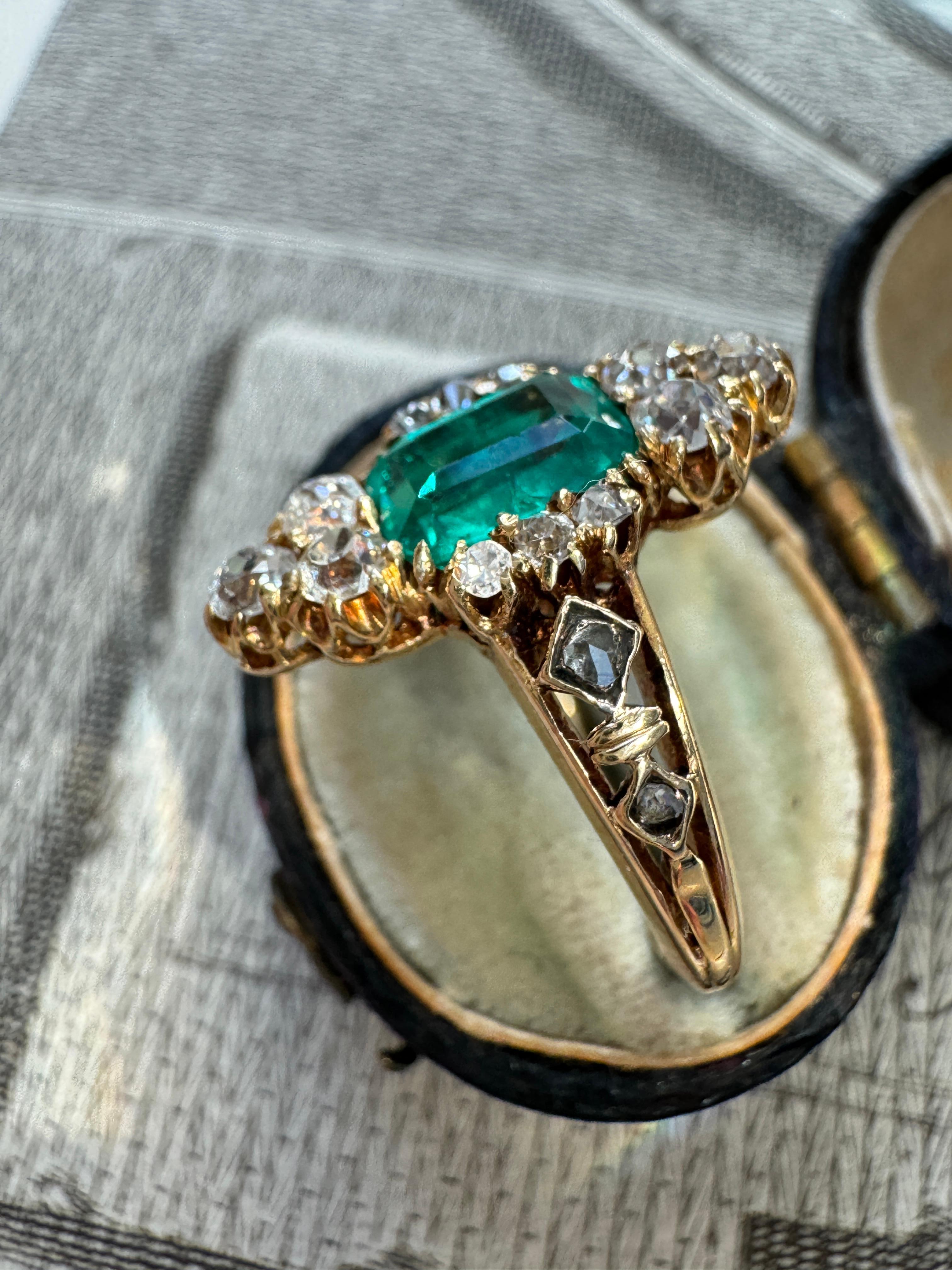 Ein leuchtender kolumbianischer Smaragd mit einem Gewicht von knapp einem Karat leuchtet zwischen einem Paar funkelnder Diamanten im Altminenschliff, die von dekorativen Diamanten im Rosenschliff hervorgehoben werden. Dieser reizvolle