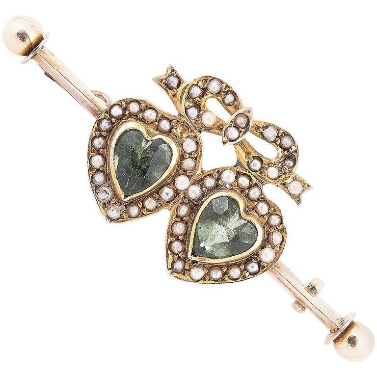 
A Victorian gold dress pin decorated with two heart shaped green peridots framed by a gold and micro seed pearls knotted ribbon, overall length 43 mm.
