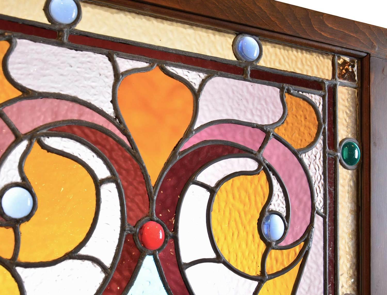 Made circa 1895, this vibrant stained glass window has Victorian scrolling, stylized fleur-de-lis, a beveled center, and ten round red and blue jewels incorporated into the design. The amber glass border has another 18 blue and green jewels with