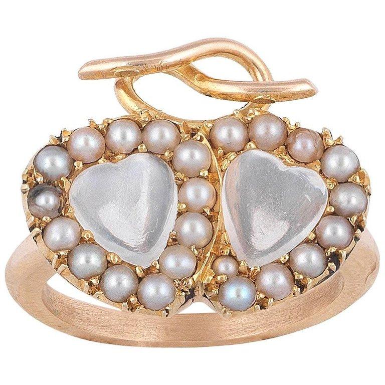 A late Victorian moonstone and pearl twin heart ring, the ring comprising two interlocking hearts, each set to the centre with a moonstone to a perl border, with gold ribbow surmount, all grain-set to a yellow gold mount and tapered D-section shank,
