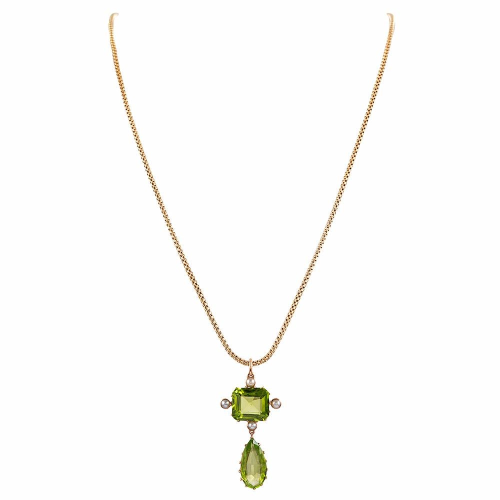 A cushion of peridot is speckled at its compass points with a solitary seed pearl and suspended therefrom is an additional pear peridot. The pendant is rendered in 9 karat yellow gold and likely was made in the United Kingdom. The 16 inch chain is