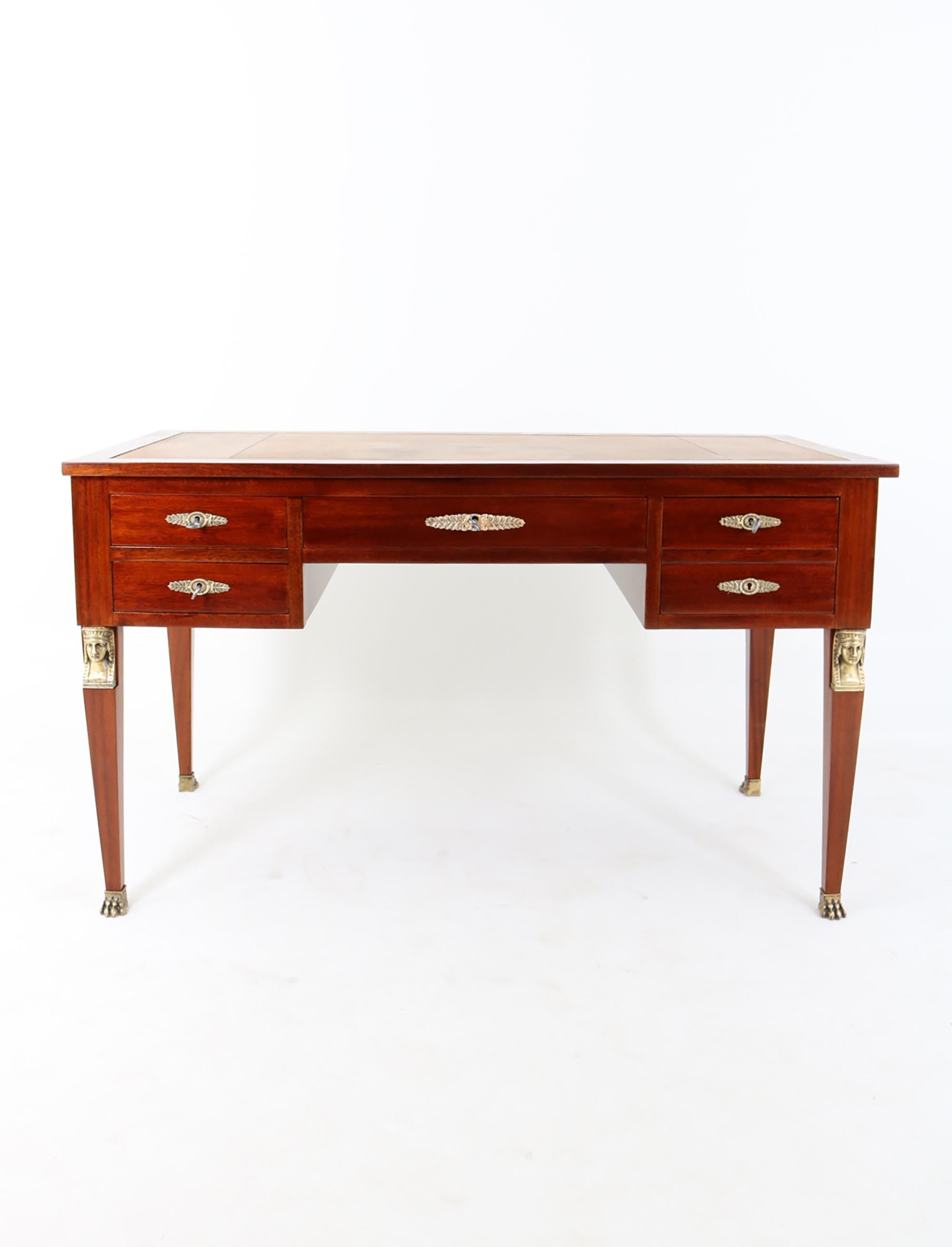 Empire desk with mahogany veneer, decorated on both sides with brass applications in the form of caryatids and floral motifs. The piece of furniture is equipped with functional lifts, which, like the top, are covered with leather and decorated
