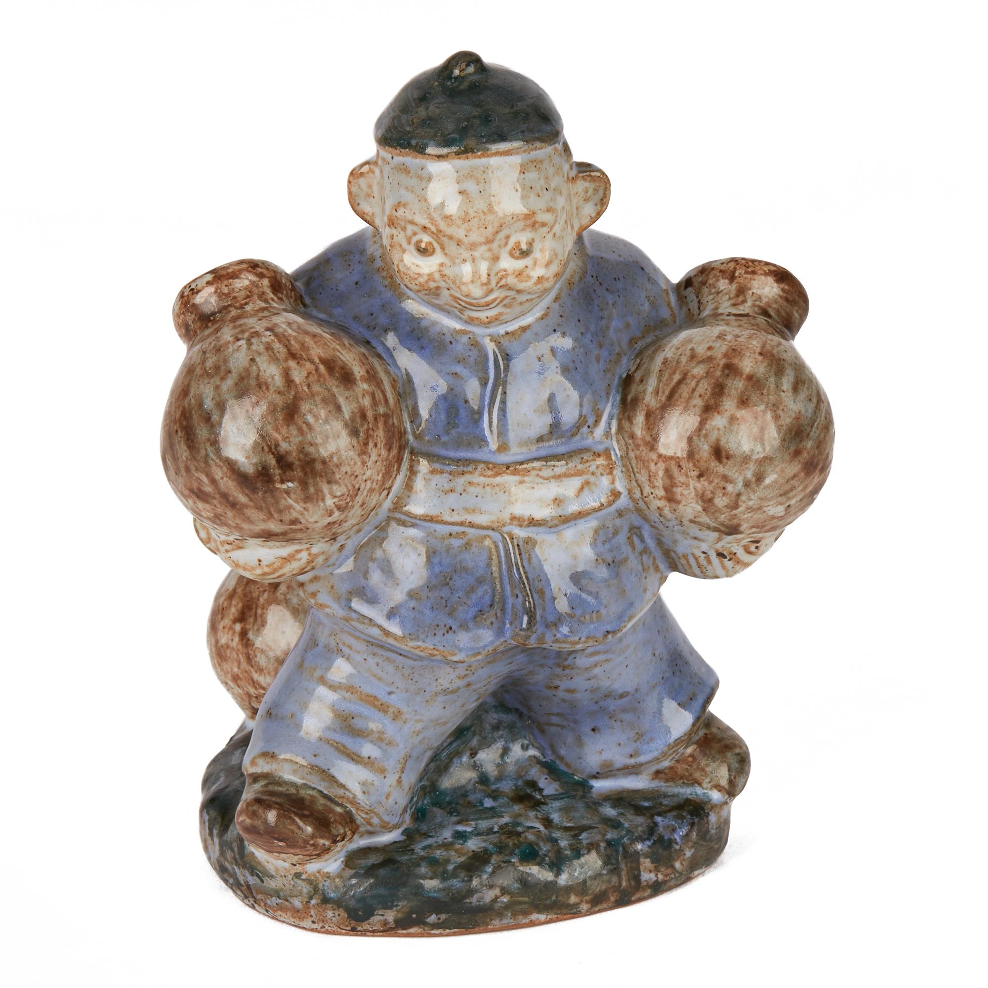 A stylish midcentury Danish stoneware pottery sculpture of an Asian figure carrying two large pottery vessels with further vessels standing on the base by L Hjorth. The large figure is glazed in blue and brown on a grey colored ground and is mounted
