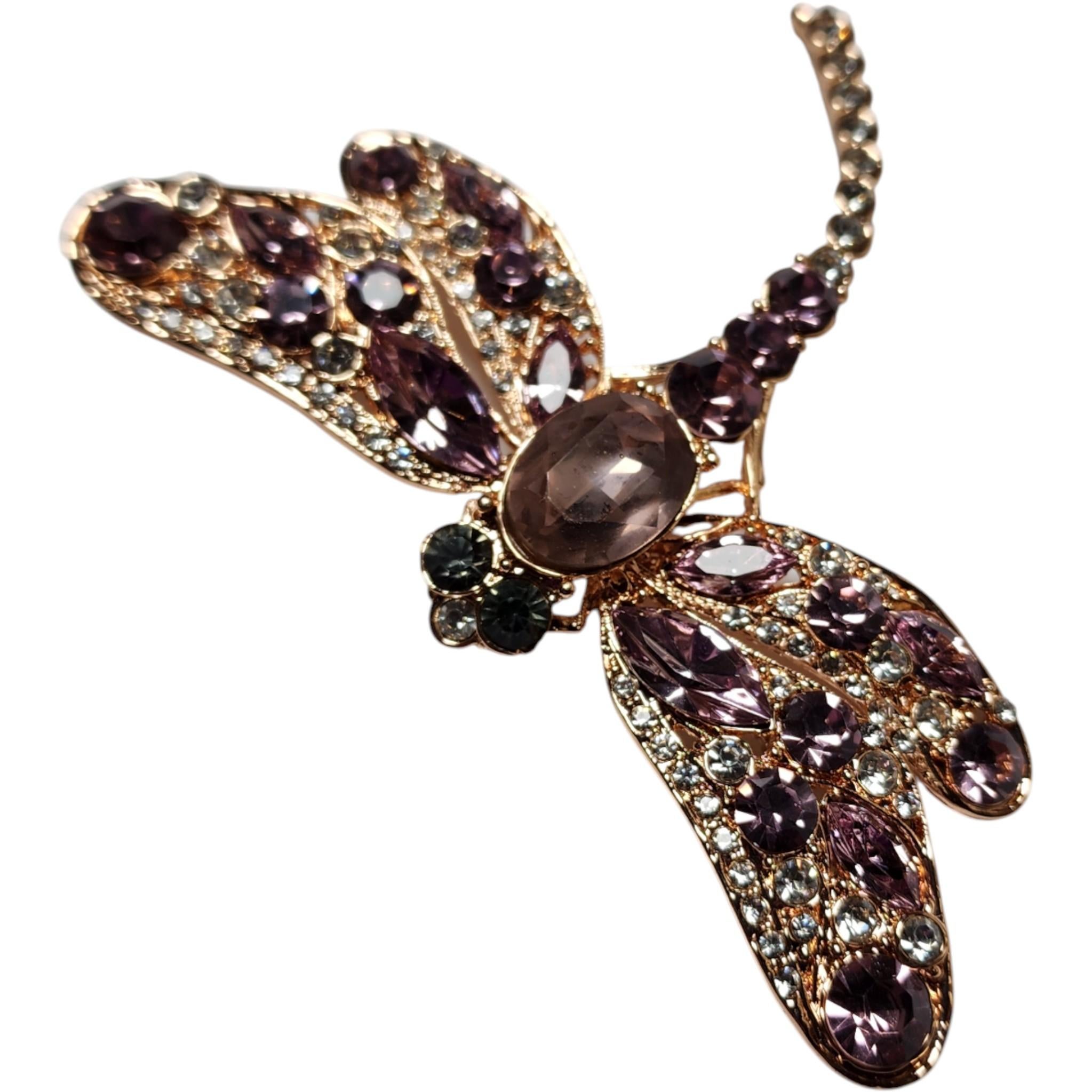 Questa incantevole spilla a forma di libellula abbaglia con strass color lavanda, accentuati da scintillanti cristalli chiari e da un sottile tocco di verde fumé sugli occhi. Le sue ali sono meticolosamente incastonate con un mix di pietre rotonde,