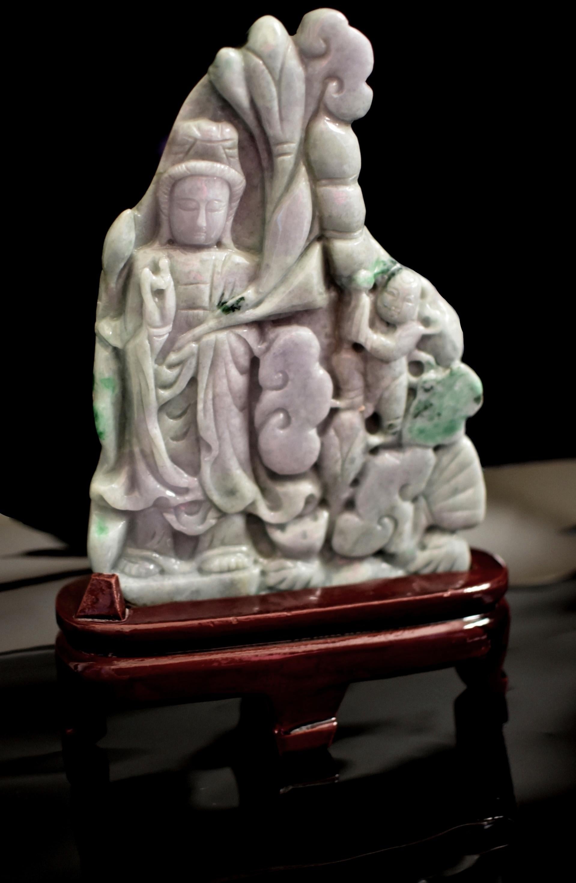 Magnifique statue en jadéite massive, sculptée à la main, représentant Kwan Yin et son fils. Kwan Yin a un visage entier avec des sourcils arqués et des yeux baissés qui dégagent une aura sereine. Elle porte une longue robe fluide avec une capuche