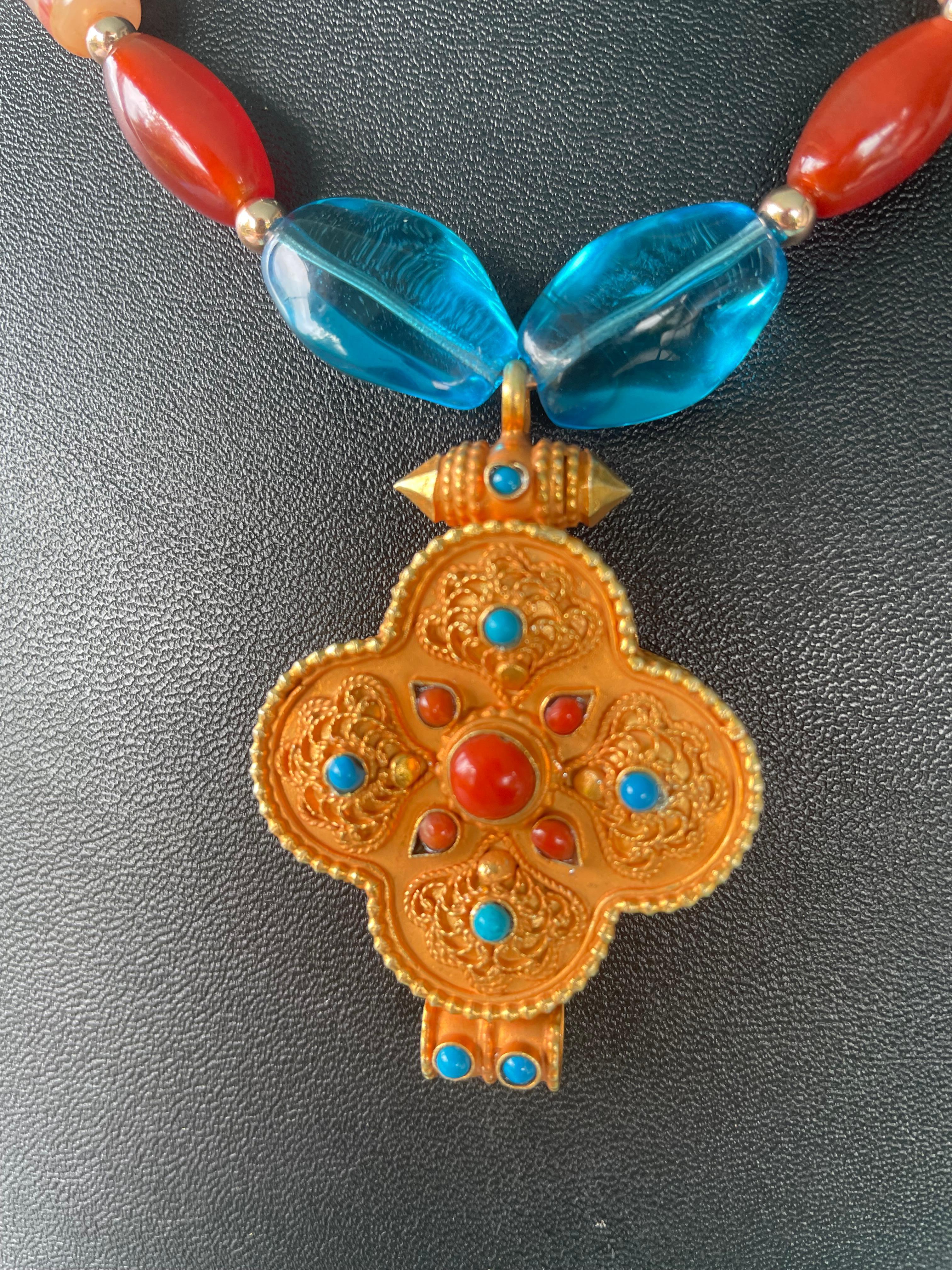 Lorraines' Bijoux propone un ciondolo Vermeil (oro su argento 925) a forma di trifoglio intarsiato, con corallo e turchese, proveniente dal Nepal. Il vetro blu vintage e le perline veneziane aggiungono un contrasto alle deliziose perline di agata e