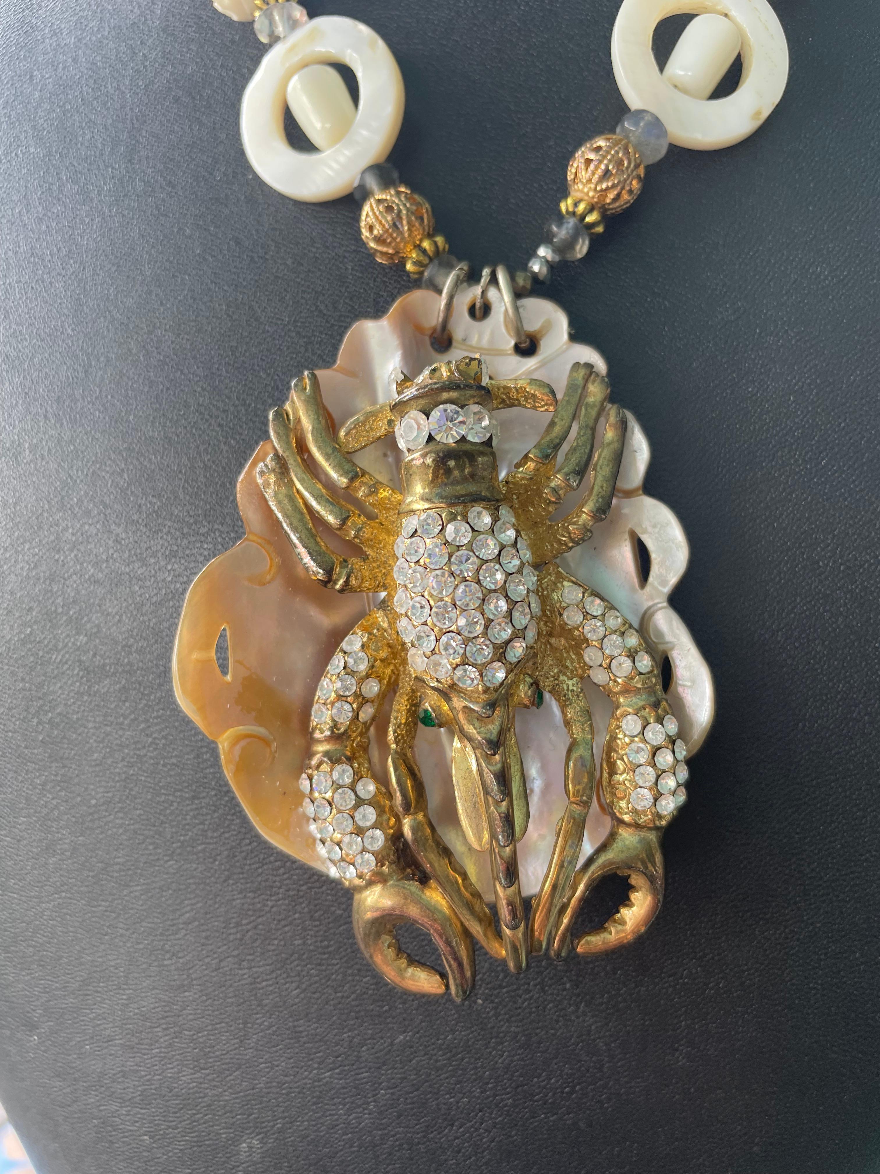 Lorraines' Bijoux propone un ciondolo a forma di conchiglia smerlata in madreperla e una grande spilla aragosta vintage anni '60, in oro e cristallo, con occhi in cristallo verde. Le perline sono in osso intagliato, madreperla intagliata, accenti