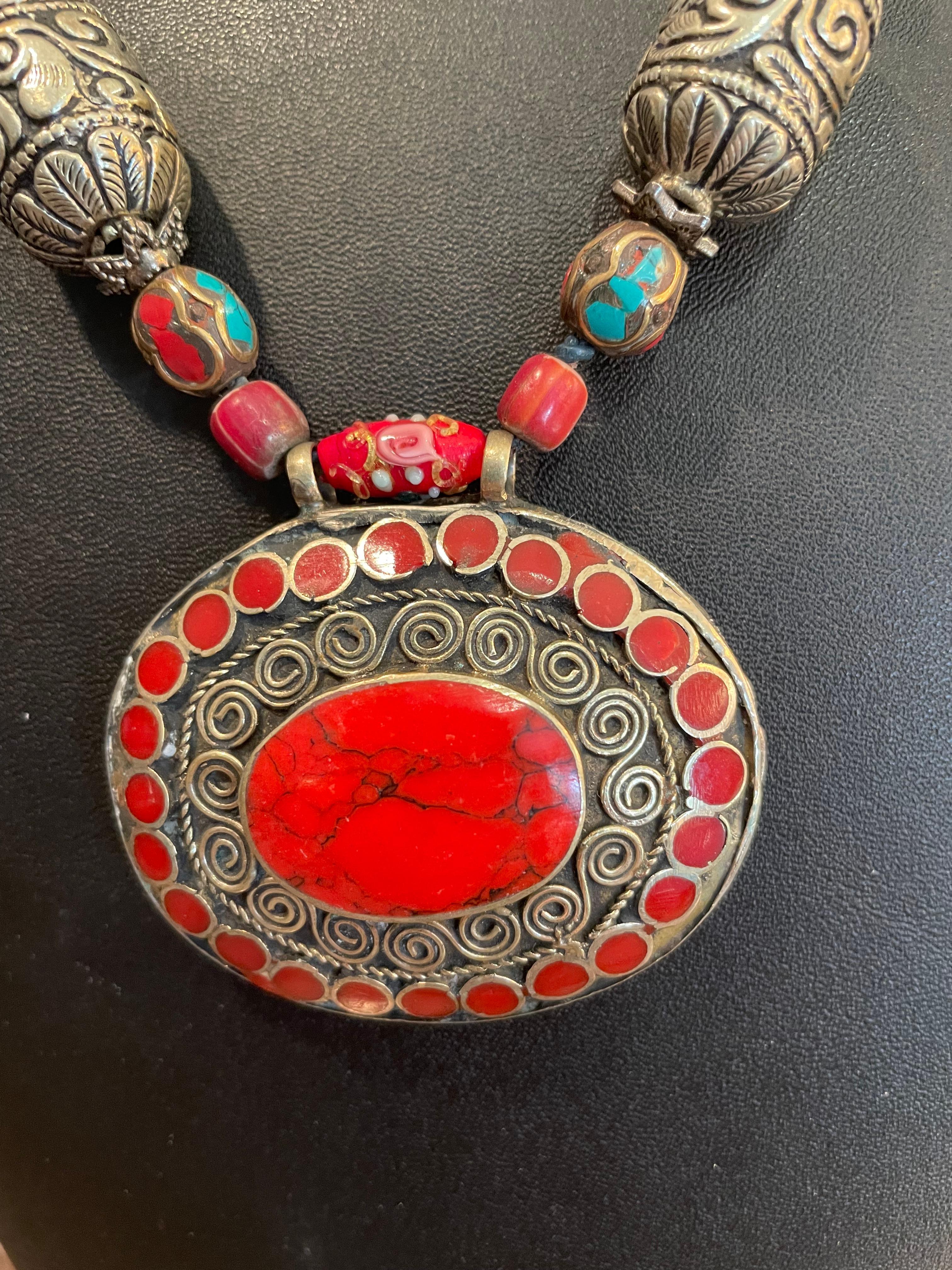 LB propose un collier pendentif tribal unique, fait à la main. Les perles vénitiennes en verre rouge rehaussent la pierre centrale rouge et les pierres rouges qui l'entourent, tandis que les grosses perles en argent tibétain ajoutent de la richesse
