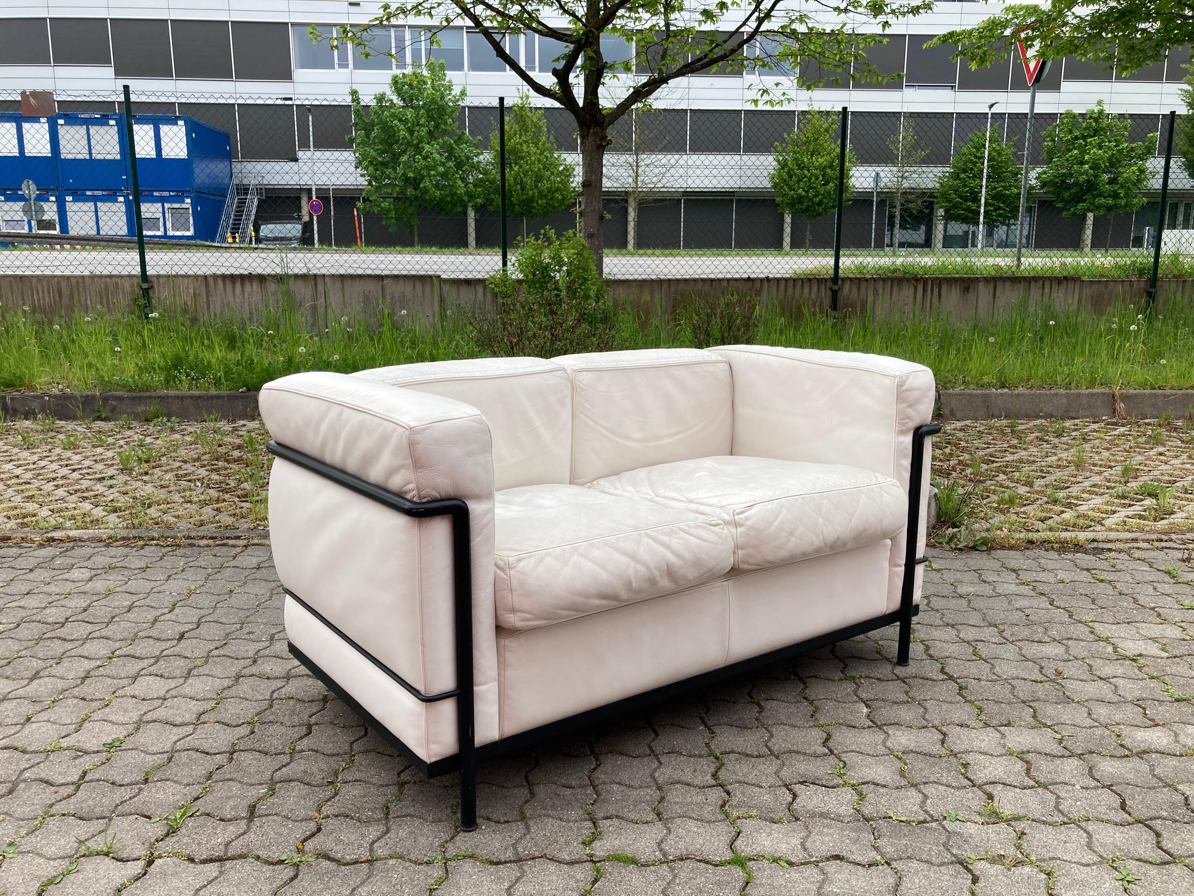 This LC2 two seater sofa in white leather was designed by Le Corbusier and produced by Cassina.
It features a black colored tubular steel frame. 
The leather has some patina.
Some marks on the tubular frame.
A Classic Iconic Sofa in a beautiful