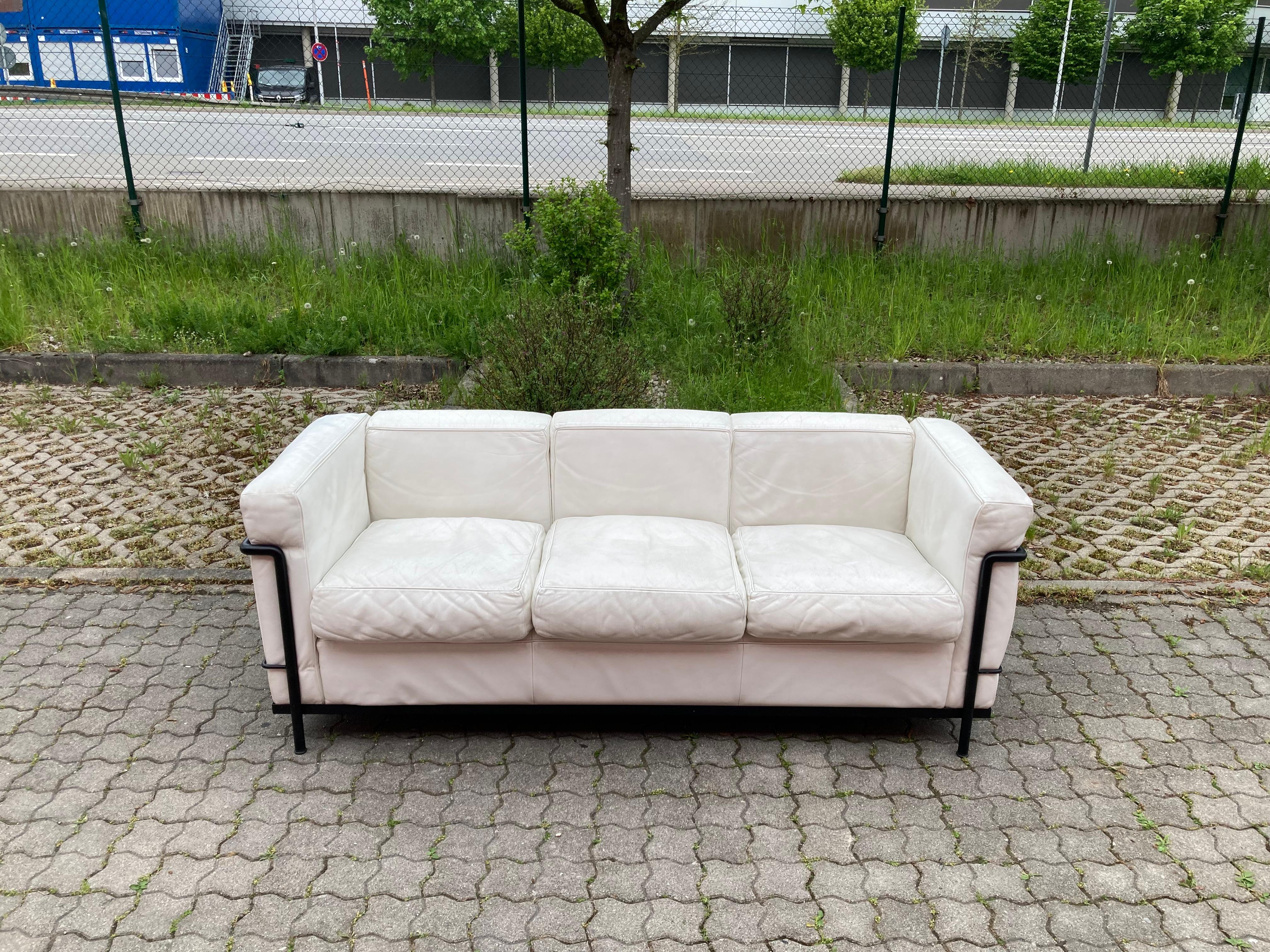 This LC2 three-seat sofa in white leather was designed by Le Corbusier and produced by Cassina.
It features a black colored tubular steel frame. 
The leather has some patina.
Some marks on the tubular frame.
A Classic Iconic Sofa in a beautiful