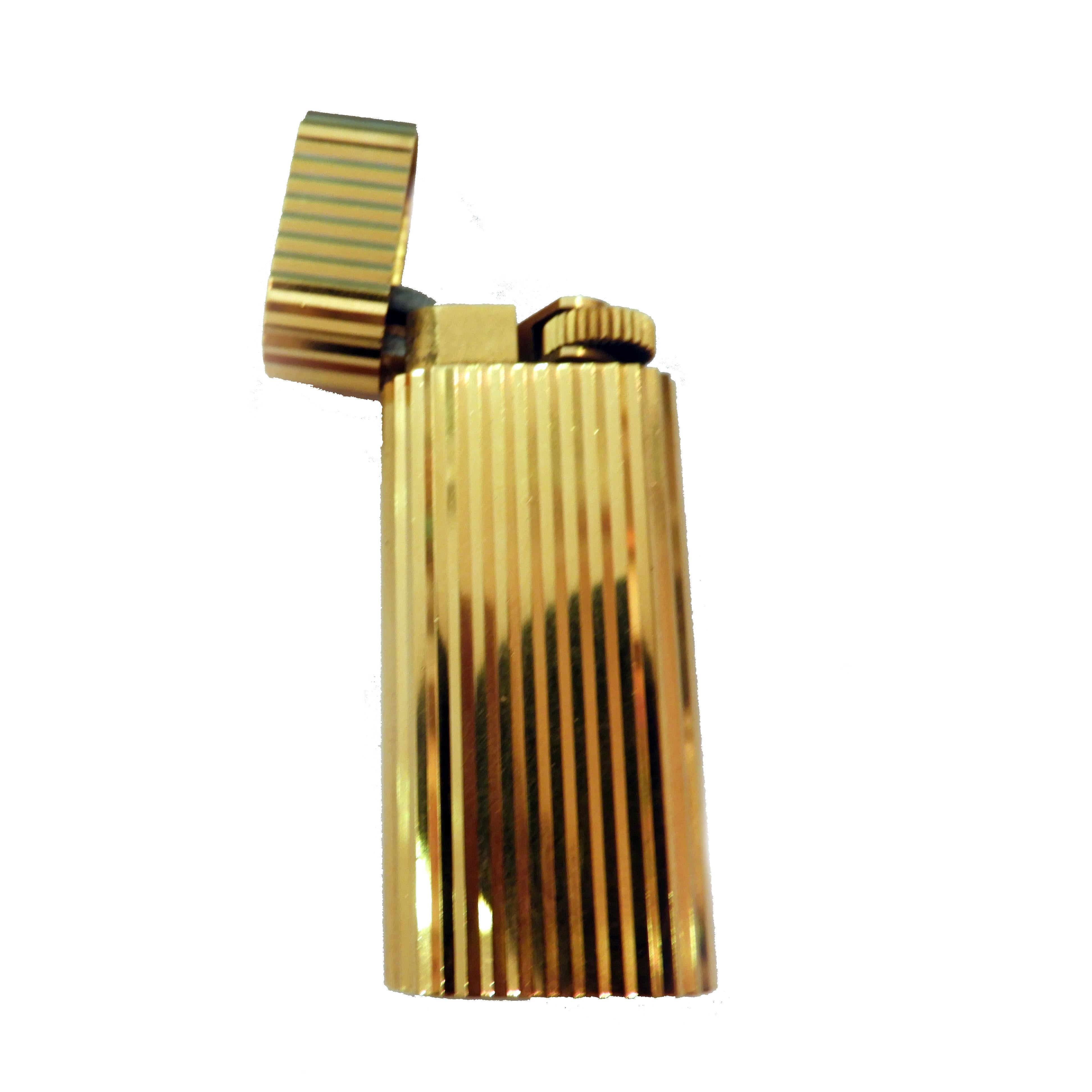 Pocket Le Must de Cartier lighter  in 18-karat gold-plated textured finish a ribbed/fluted design. 
It has a flip-top lid that houses the Swiss-made mechanism. The flint wheel pivots when opened and closed as its designed to be offset and exposed