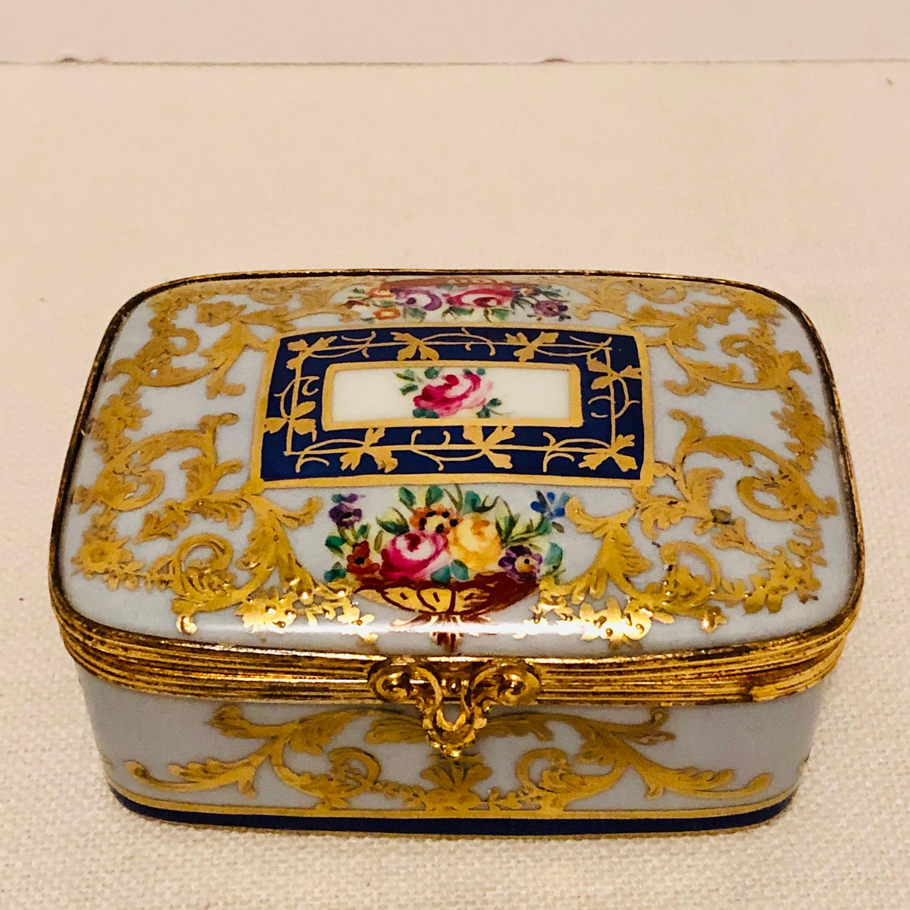 This is an exquisite Le Tallec porcelain box with a light blue background color. It is decorated with two flower bouquets and an arabesque of intricate raised gilding on every side. It has an accent of a cobalt colored square with raised gilding in