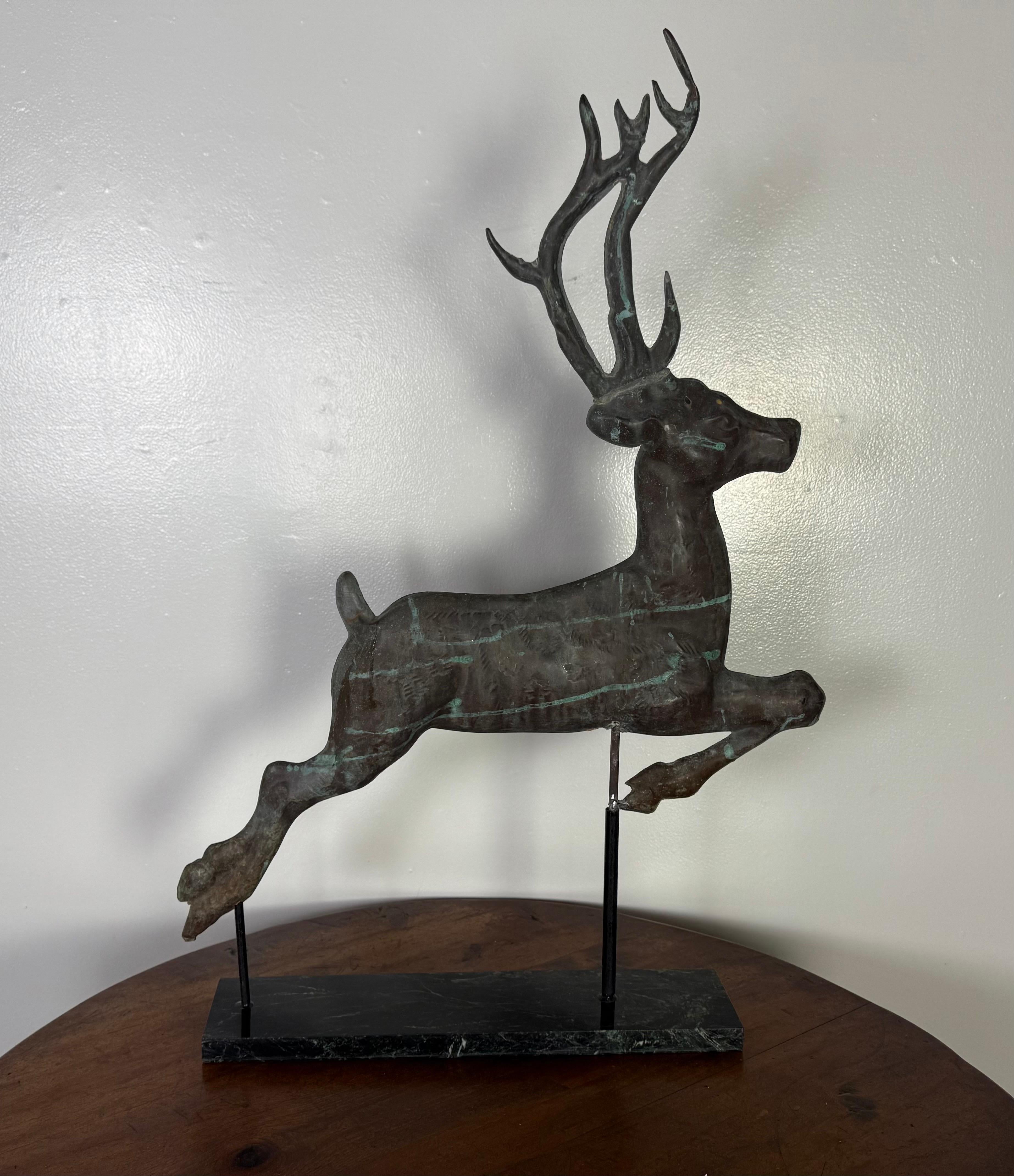 Vintage leaping stag weathervane made of copper, with a verdigris patina that gives it a weathered, greenish appearance.   Then stag is depicted mid-leap with its from legs extended forward and hind legs stretched back, conveying a strong sense of