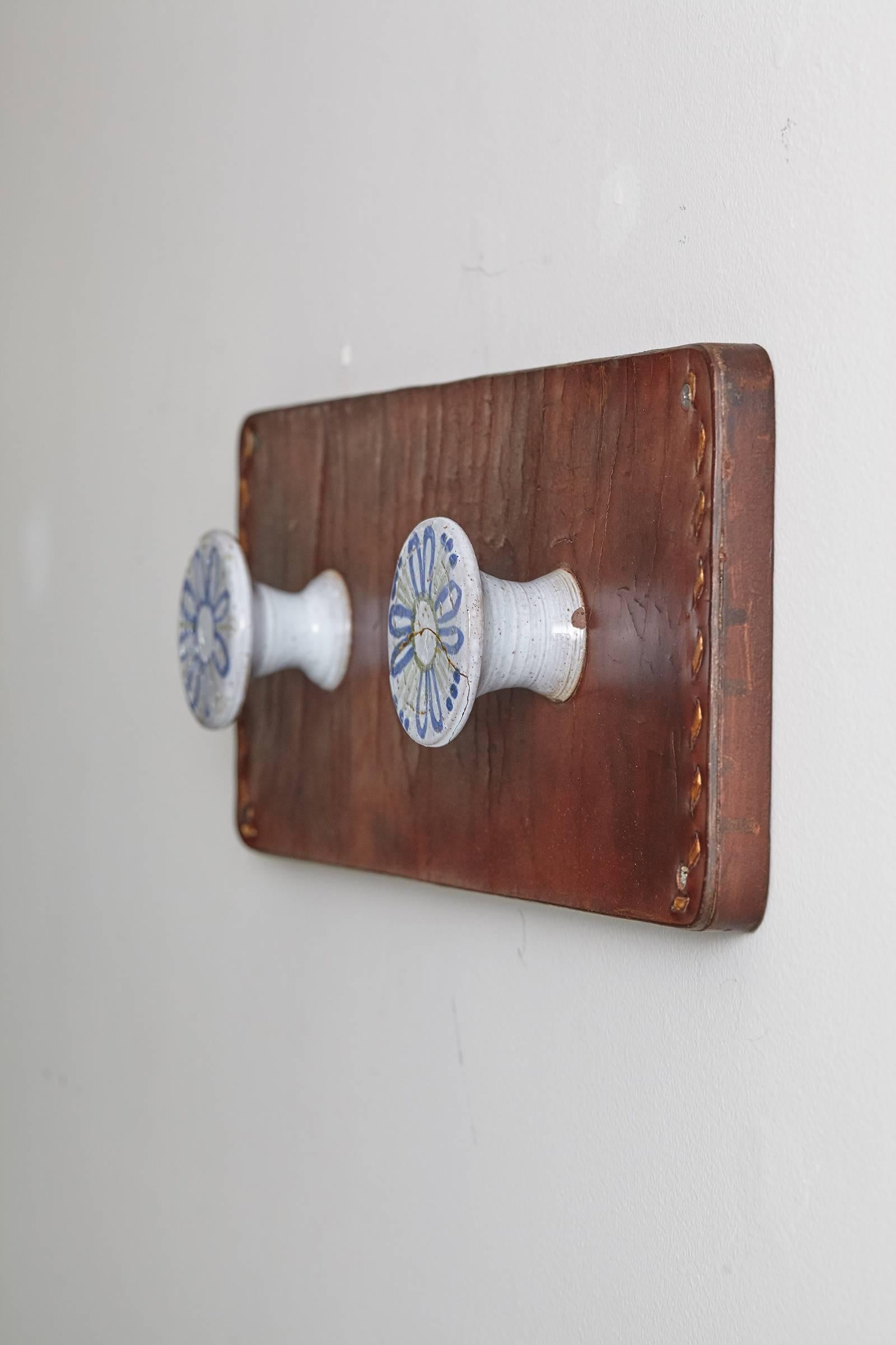 Beautiful and unique wall hook made of leather with stitching detail and hand-painted ceramic knobs. One knob has been cracked and repaired but still in gorgeous vintage condition. Great for hats and entryway necessities.