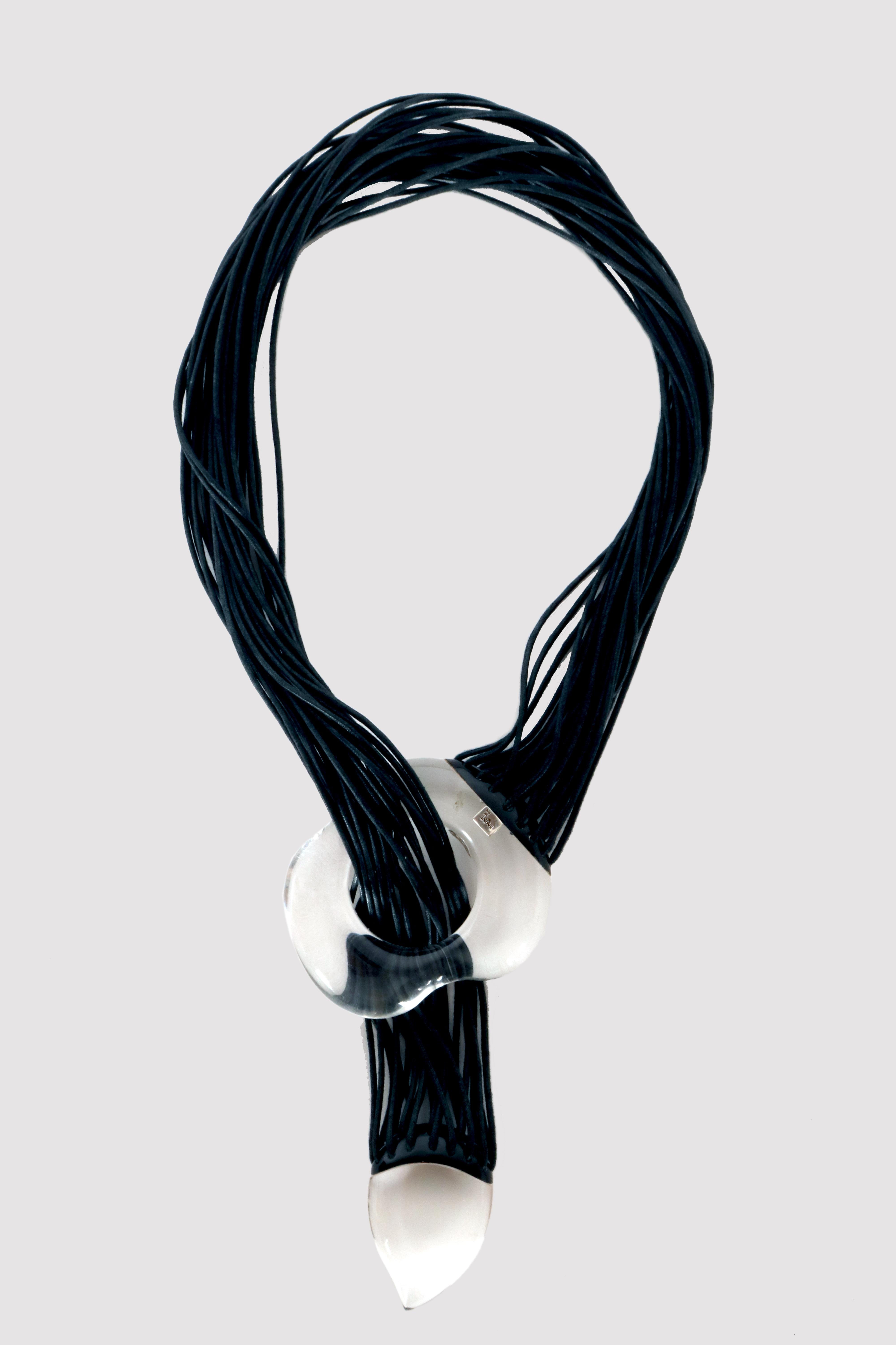 Collier de fantaisie composé de 27 minces fils de cuir noir, attachés aux deux extrémités à deux éléments en lucite noire et transparente, l'un percé et l'autre en forme de goutte d'eau, aux courbes organiques. Poinçon de fabrication en argent