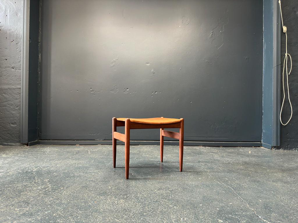 Questo poggiapiedi compatto in pelle è un pezzo eccezionale. Con un design semplicistico, caratterizzato da gambe affusolate in teak con dettagli rotondi intagliati nella parte superiore della struttura. Grazie alla seduta imbottita in pelle