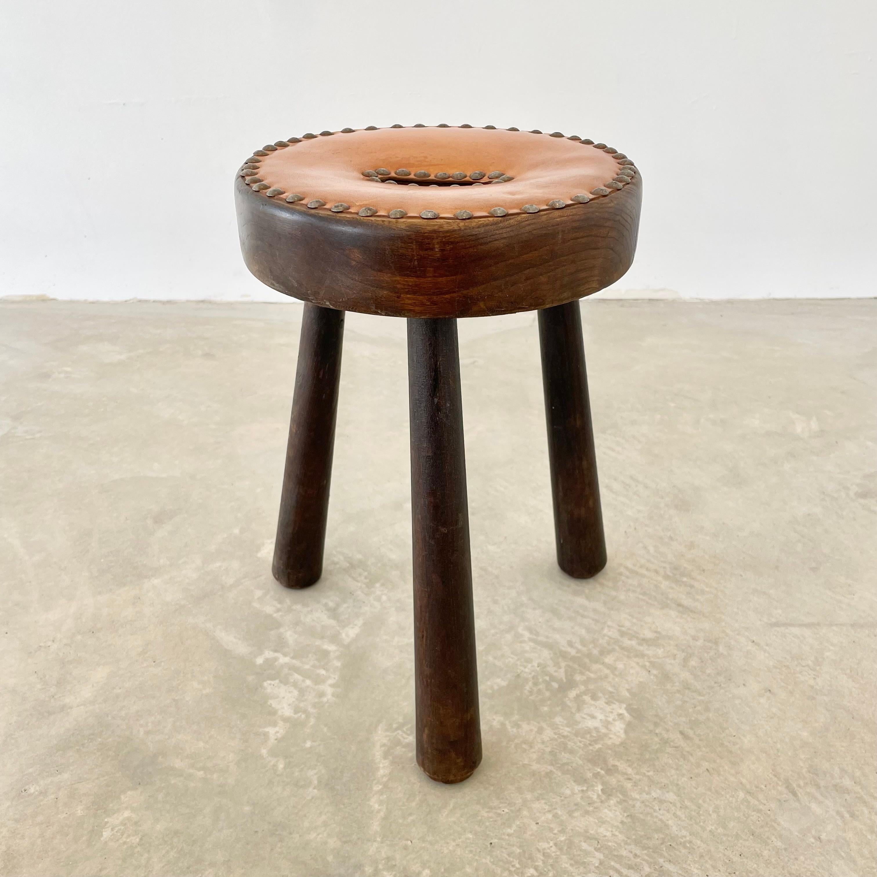 Unique tripod stool made in France, circa 1970s. Substantial and chunky round seat with three sturdy club legs that taper out at the bottom. Seat is covered with a thick piece of leather that is studded into the wood with metal studs all around its
