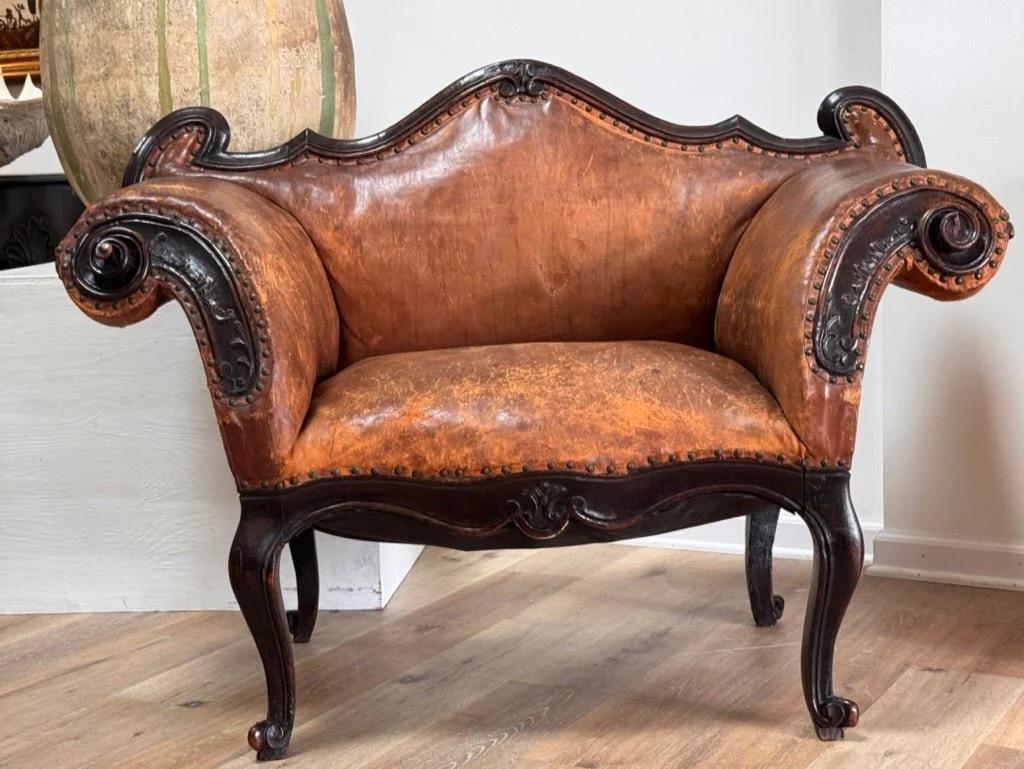 Italian Lombardi small wide chair or banquette or marquis with deeply carved  “rams head” armrests, mid 18th century. Having early, possibly original leather. 38.25h. x 35” w. x 27.5” d. SH 20

