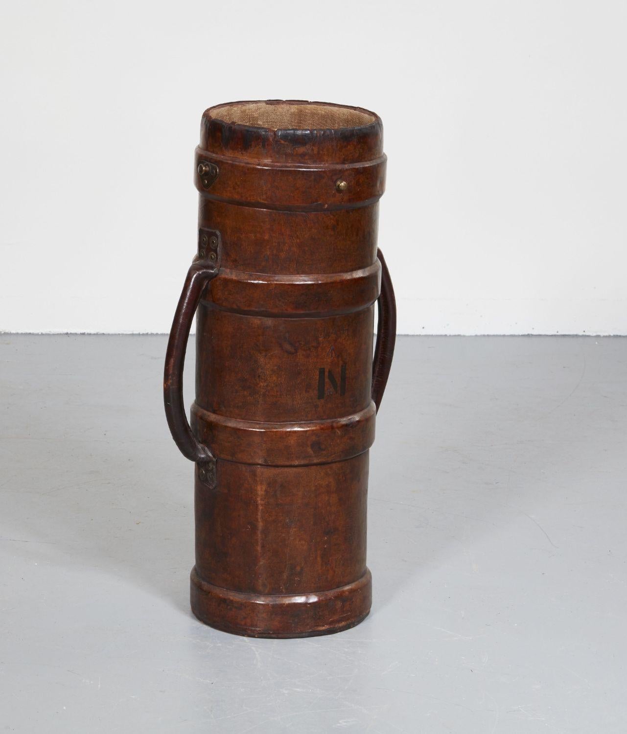A tall military powder cartridge carrier in polished leather having ribbed sections and two riveted side-mounted carrying handles also in leather. Embossed JAH and N. Useful as an umbrella stand or holder for cherry blossoms and other decorative