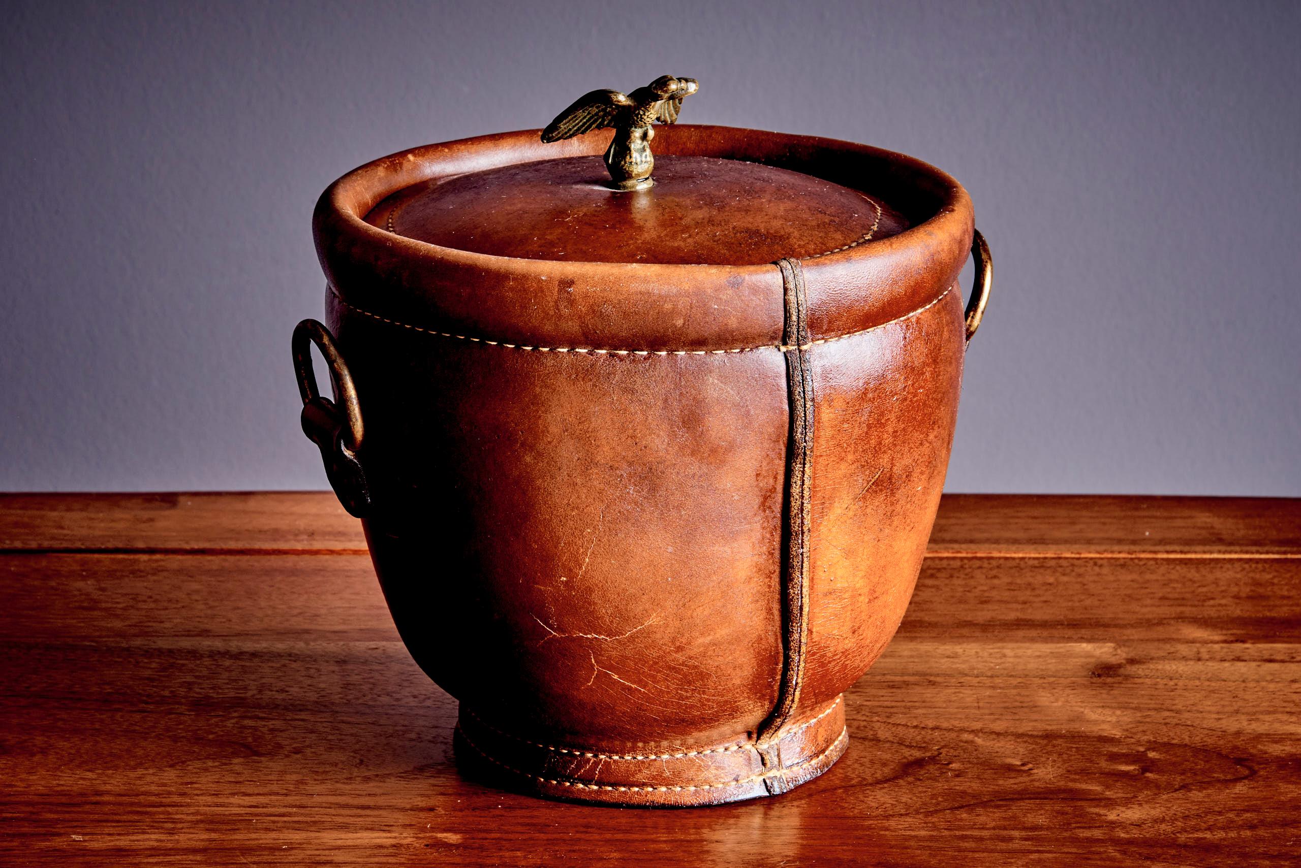 Vintage ice bucket wrapped in rich brown leather with visible stitching and a warm patina. The lid is topped with a detailed brass eagle finial, giving it a distinctive and stately look. The bucket features brass side handles and a removable