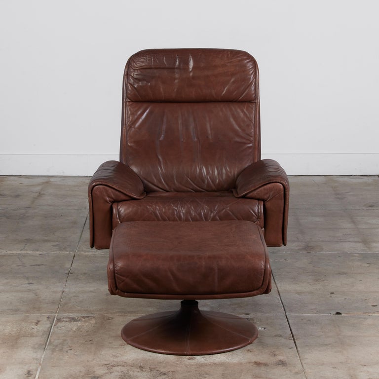 Lounge chair and ottoman for Swiss manufacturing company Artima, c.1970s. The set features a fully upholstered chair and ottoman in a patinated brown leather with stitched base detail. The chair has two clever pockets on each side to hold magazines