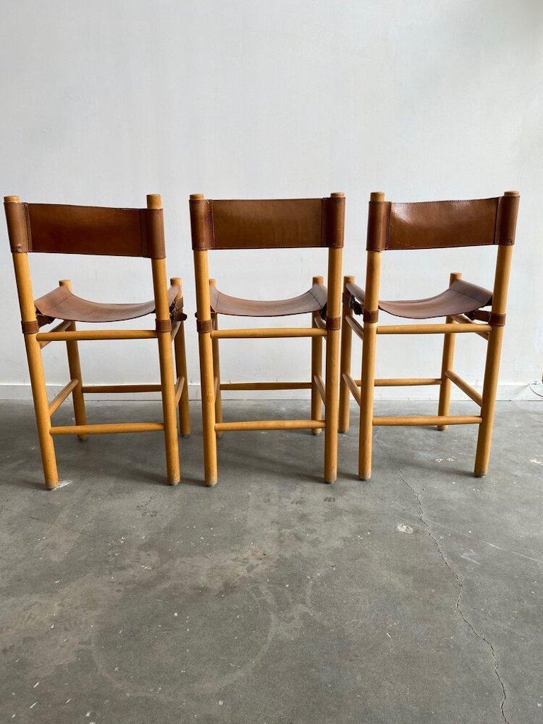 A set of three brown leather safari mid-century barstools in the manner of Michel Arnoult. These counter-height stools feature heavy stitched leather, side buckle detailing, and premium soft leather safari-style seats and back. “Made in Argentina”.