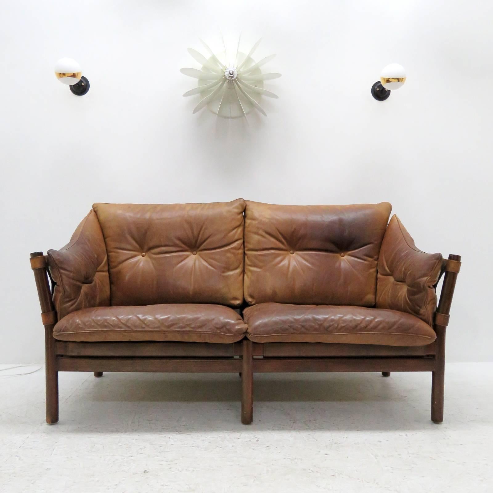Stunning leather settee designed by Arne Norell in the early 1960s and produced by Norell Möbel AB, Sweden, with thick tufted leather cushions on saddle leather sling supports with brass hardware, the frame is made of sturdy, dark stained solid